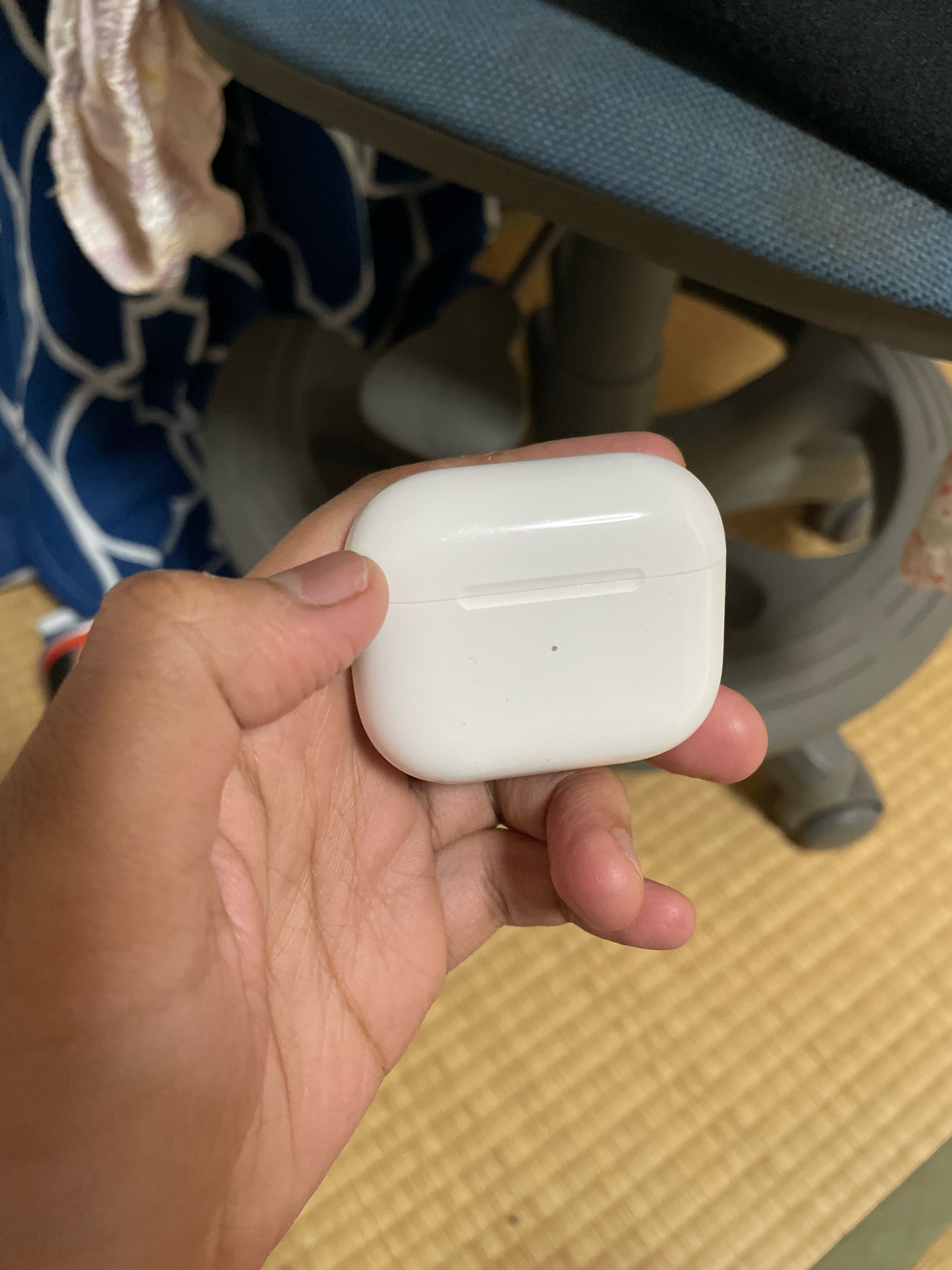 A person's hand is holding a white charging case for wireless earbuds. The case is oblong with rounded edges and has a subtle seam running across its top. A small dark dot is visible on the case's surface. The background features a tatami mat floor, part of a chair with a blue textured fabric seat, and a blurry dark object with a metallic sheen. The scene appears to be indoors, possibly in a home or office environment in Yokohama, Japan, given the tatami mat flooring which is a traditional Japanese flooring material.