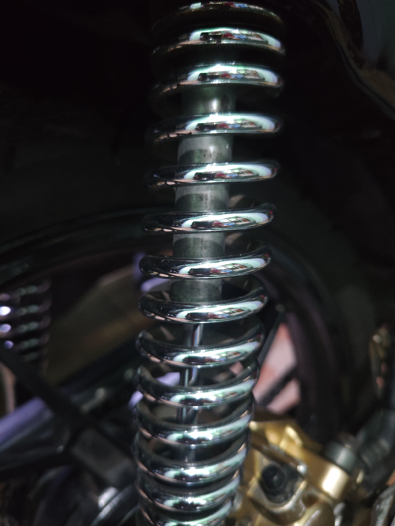 This is a close-up, low-angle, vertical shot of a motorcycle's rear shock absorber, focused on the chrome spring. The shock absorber is positioned on the right side of the frame, with its coiled spring dominating the center. The spring is a shiny, metallic chrome, reflecting ambient light and colors, creating a pattern of highlights and shadows along its coils. Behind the shock absorber, the background is dark and out of focus, with hints of black plastic, metal components, and some blurred reddish and golden elements suggesting other parts of the motorcycle. The scene appears to be in a dimly lit environment, possibly a garage or workshop in Sroyo, Indonesia, where the motorcycle is being examined or worked on. There are no people visible in the image.