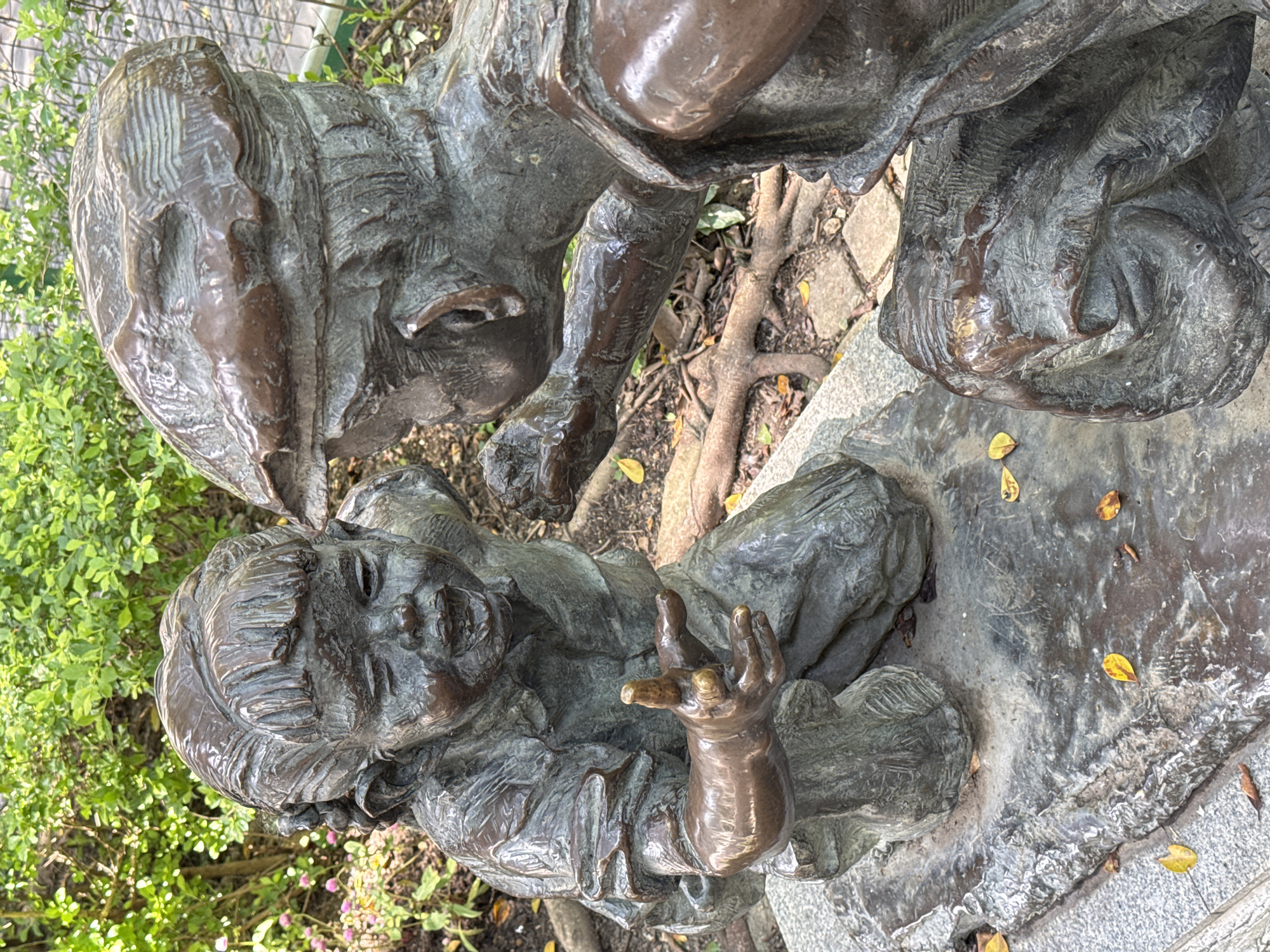 The image shows a bronze statue of two children. One child is lying down, looking up with a hand raised. The other child is looking down at the child on the ground. The statue is located outdoors, near greenery and a stone path. Based on the location information provided in the prompt, the statue is located in Liwan District, China.