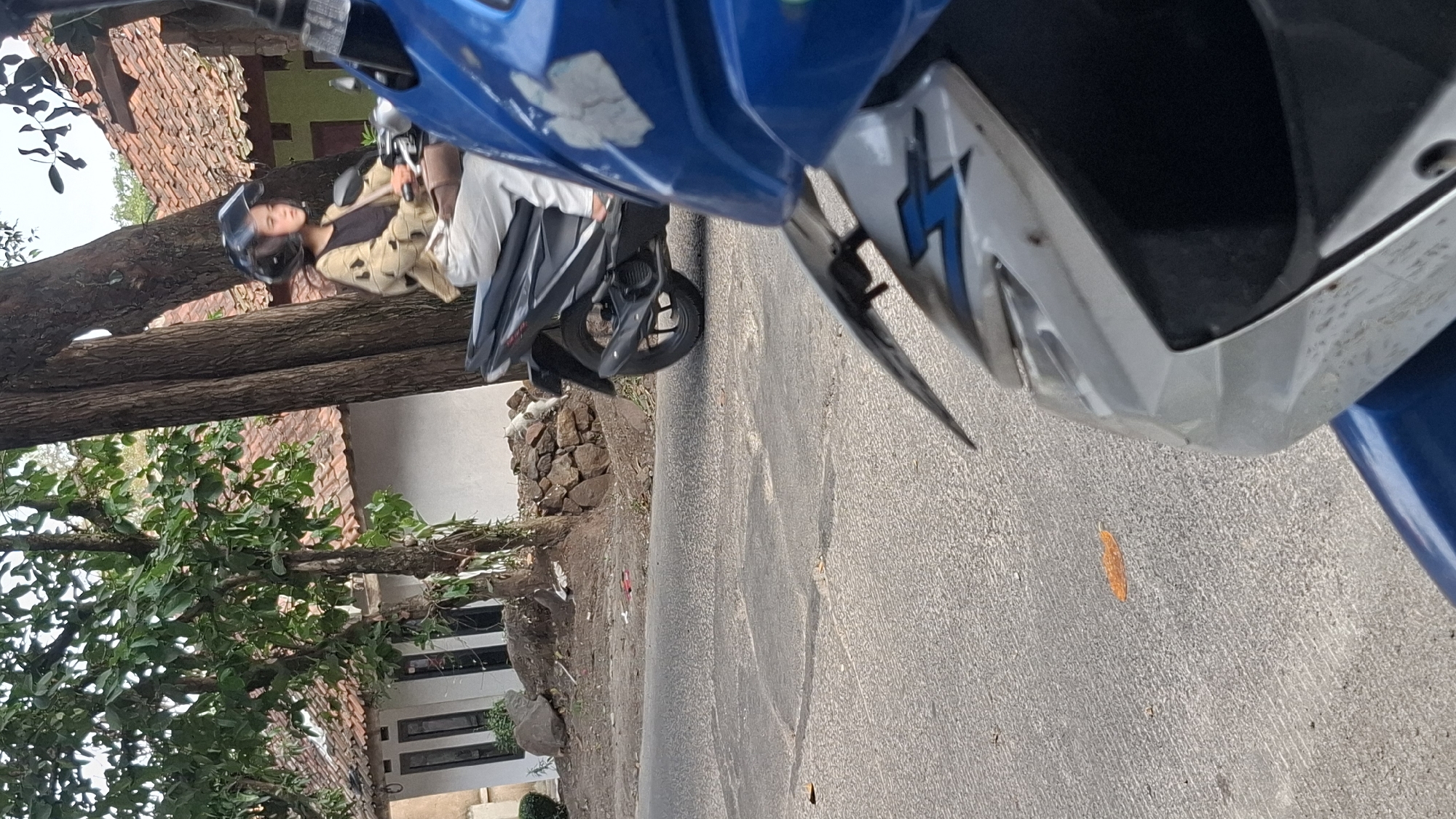 In Cilaku, Indonesia, a young woman wearing a helmet and a patterned cardigan over a dark top rides a black motorcycle. She is looking to her left. The motorcycle is parked on a paved road. In the background, a house with a red tile roof and white walls is visible, surrounded by trees. The scene suggests a casual, everyday moment in a residential area.