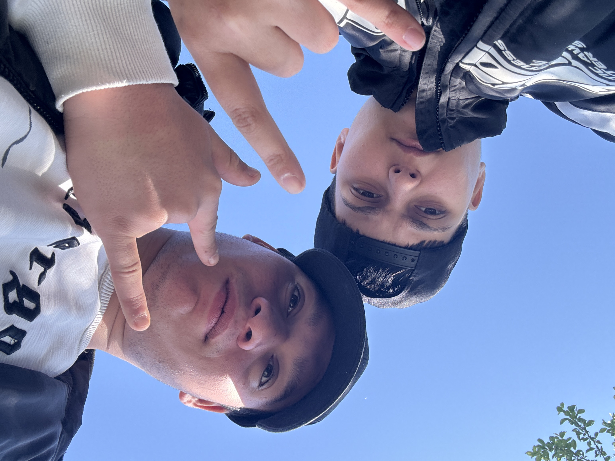 This media file presents an upward-angled view of two young men, likely taken from a low perspective looking up at them. The setting is outdoors, characterized by a clear, bright blue sky that dominates the background, suggesting a sunny daytime.
The individual on the left, from the viewer's perspective, wears a white long-sleeved top with black stylized text partially visible, and a dark outer garment or vest. A dark baseball cap is worn backward on their head. Their right hand is raised, with the index and middle fingers extended in a gesture towards the camera.
The person on the right is dressed in a black jacket featuring white graphics or text, including the word "PRESS" and partial letters that might be "AW" above it. They also wear a dark baseball cap backward. Their left hand is raised, with the index finger extended. Both individuals have neutral expressions and are looking down towards the camera.
Sunlight clearly illuminates their faces, emphasizing the bright weather conditions. In the bottom right corner of the image, a small portion of green foliage is visible, further confirming the outdoor environment. The interaction appears to be a posed photograph, possibly a selfie.