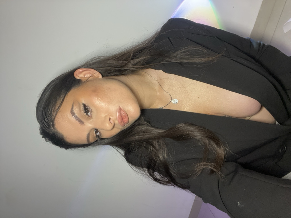 A female subject is depicted from the mid-torso upwards in a close-up portrait. She has long, dark hair, parted on the side, cascading over her shoulders. Her face is made up with eyeliner, contouring, and glossy, pouted lips, and she gazes directly at the camera. She is wearing an open, unbuttoned black blazer or jacket, which reveals her cleavage. A thin, silver-colored necklace with a small, square-shaped pendant is visible around her neck. The background consists of a plain, light-colored wall. A vertical streak displaying rainbow colors is present on the right side of the background, and a faint purple light reflection is visible on the lower left. The lighting appears bright and direct. The image does not contain any specific visual information to identify a precise location within Kiryat Bialik, Israel.
