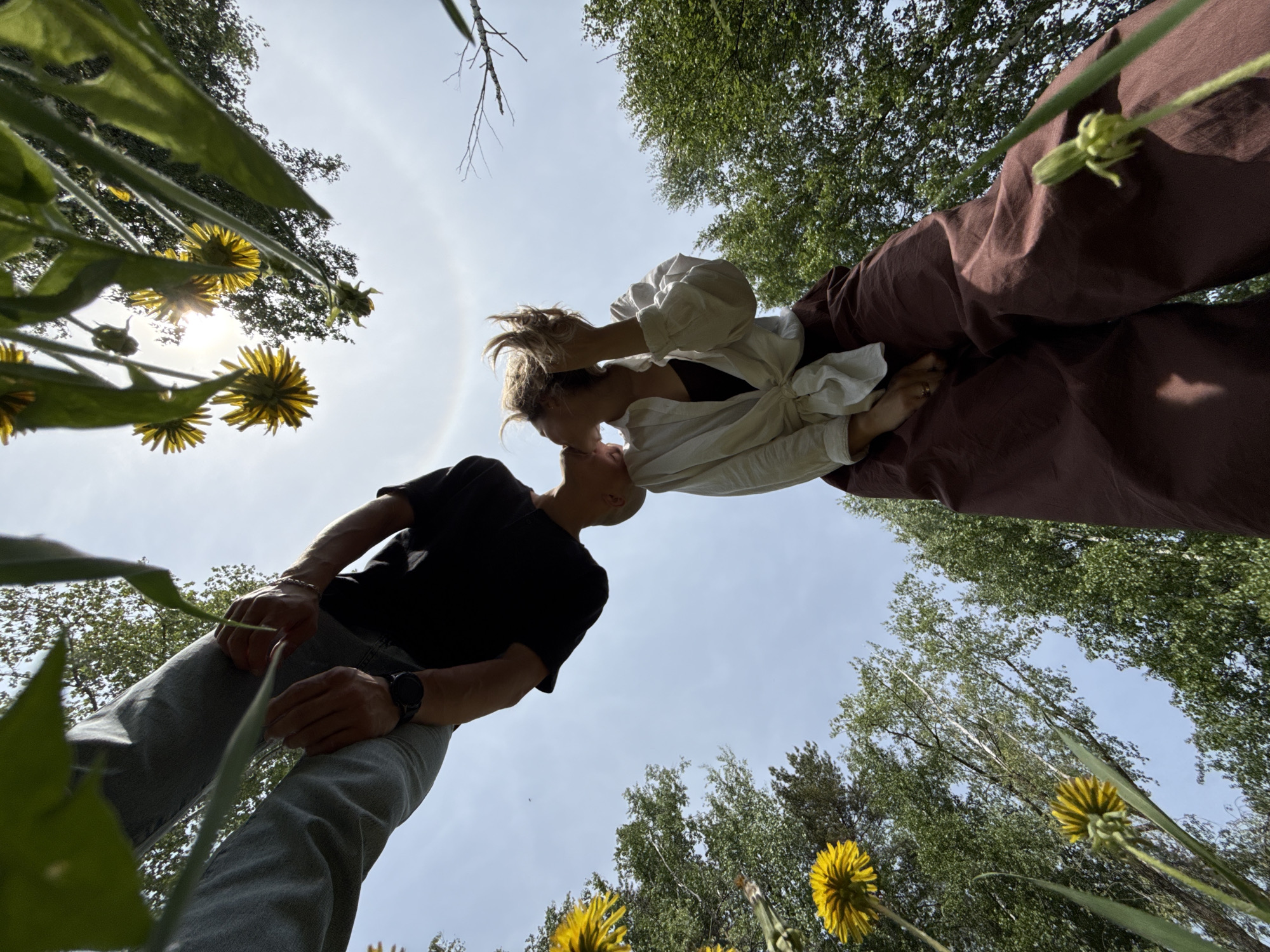 From a low-angle perspective, a couple appears to be kissing under a partially clear sky, framed by trees and dandelions. A man wears a black t-shirt, gray jeans, and a watch. A woman wears a white button-down shirt and brown pants. The location is identified as Noviy, Russia, though specific landmarks are not evident in the frame.