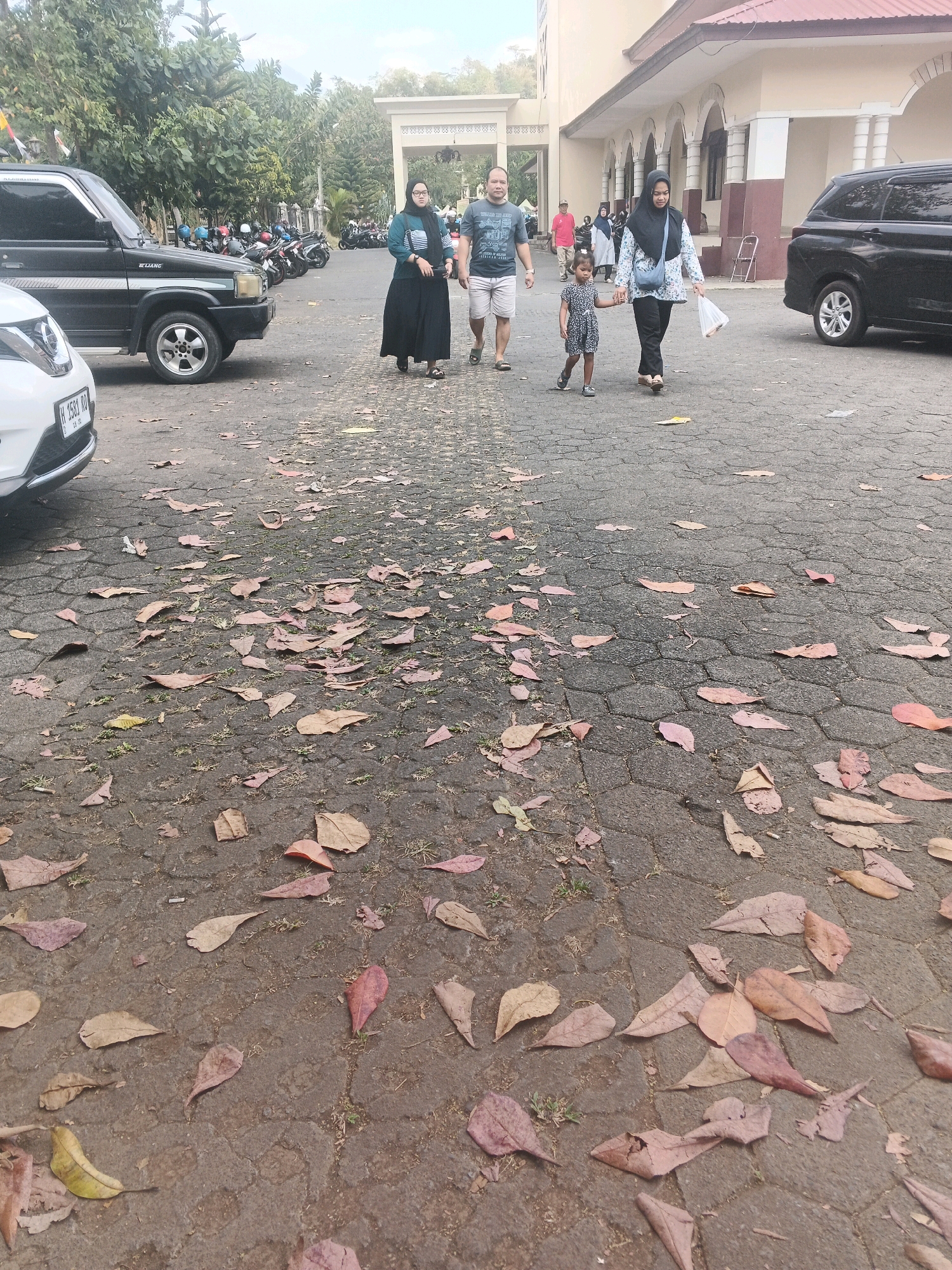 At an outdoor parking area in Kalirejo, Indonesia, a group of people walk on a paved surface littered with fallen brown leaves. To the left, a black SUV is parked. In the background, a building with arched entrances and a reddish-brown roof is visible. Several motorcycles are also parked in the background. A family, including a man, a woman in a hijab, and a young girl, walks towards the building. The man wears a dark t-shirt and shorts, the woman in the hijab wears a blue and white patterned long-sleeved shirt and dark pants, and the girl is in a patterned dress. Another woman in a hijab walks in the opposite direction on the right side of the image, holding a plastic bag. A black minivan is parked to the right of the building.