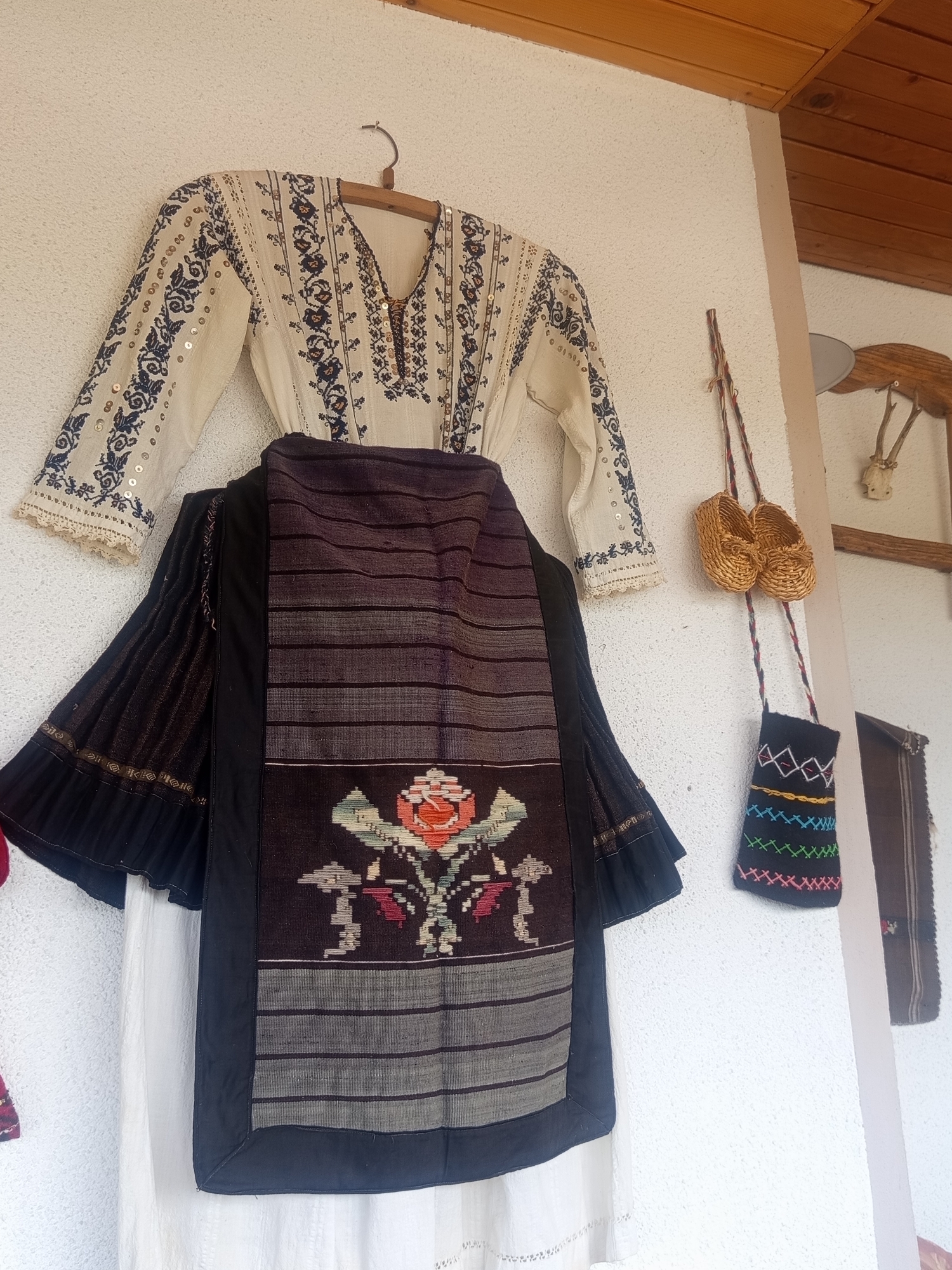 A traditional Bulgarian folk costume is displayed on a textured, off-white wall in Radishevo, Bulgaria. The ensemble consists of a long-sleeved, cream-colored blouse with a V-neckline, adorned with extensive dark blue floral and geometric embroidery and interspersed with small, metallic sequins on the sleeves and front. The cuffs feature a delicate lace trim. Below the blouse, a dark, woven textile garment, likely an apron or a decorative skirt panel, is displayed. This garment features horizontal dark grey stripes in its upper section and a prominent central embroidery of a large orange-red rose with green and grey foliage against a dark background. The edges of this garment are trimmed with plain black fabric. A white underskirt is visible beneath the dark garment. The blouse is suspended by a simple wooden hanger. To the right of the costume, a braided cord suspends two miniature, woven straw shoes (opanki) above a small, black textile bag embroidered with colorful geometric patterns in yellow, green, blue, pink, and white. In the upper right background, a wooden ceiling with horizontal planks is visible, along with a portion of a wooden rack featuring a set of antlers. A dark, vertically striped textile also hangs to the far right. The wall has a stucco-like texture.