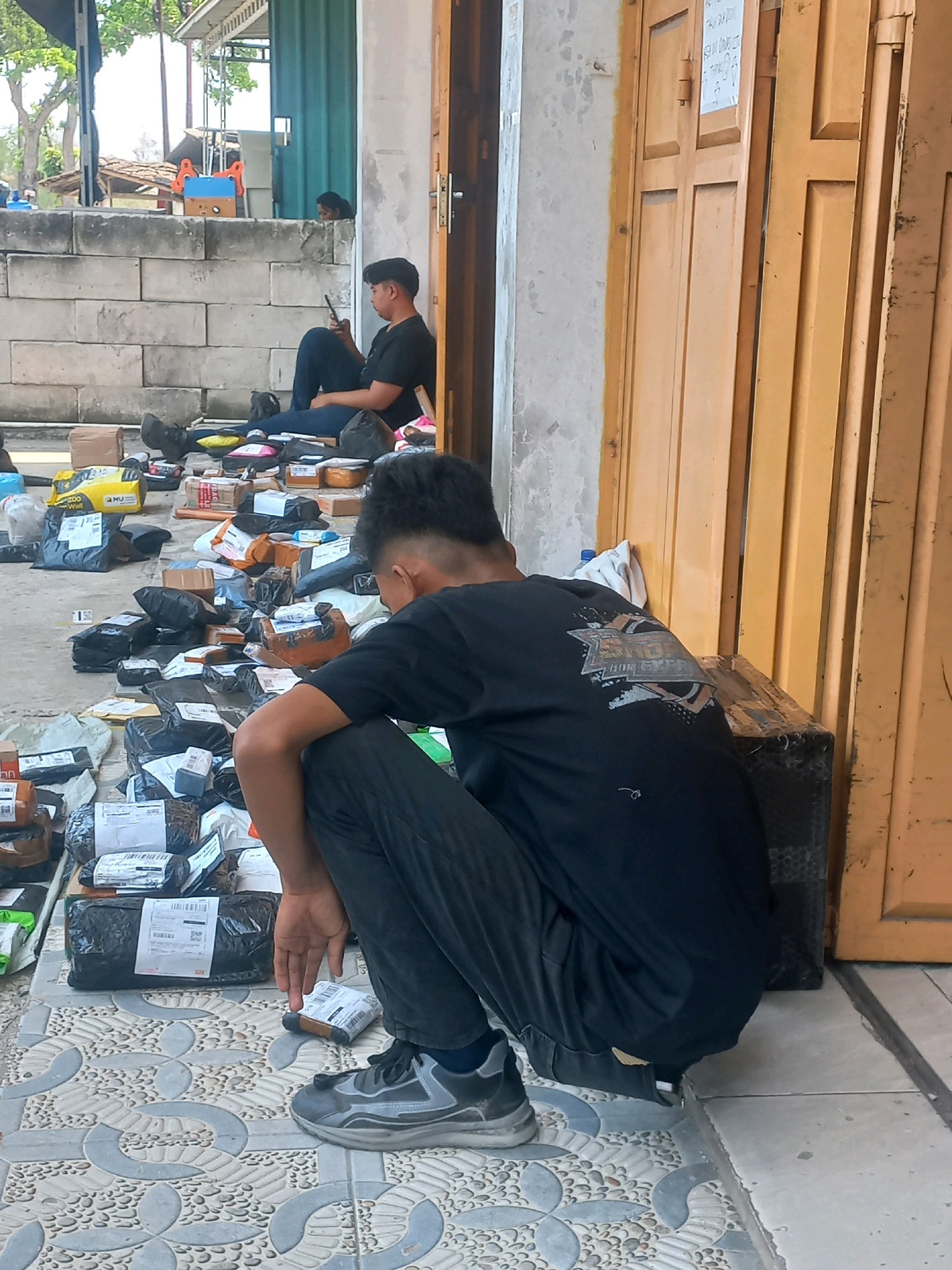 The image depicts a street scene in Baturetno, Indonesia, with a focus on a young man crouching on patterned pavement in front of an orange shuttered building. He is wearing a black t-shirt and dark pants, with grey athletic shoes. Around him, numerous packages of various sizes and shapes are scattered on the ground, some wrapped in black plastic, others in brown cardboard. To the left, a wall constructed of light-colored concrete blocks provides a backdrop. Further back, another young man is seated against the wall, looking at his phone, with more packages visible near him. The environment appears to be an outdoor area adjacent to a shop or business, suggested by the shuttered entrance and the presence of packages, implying a delivery or distribution point. Trees and a glimpse of a building structure are visible in the upper left background.