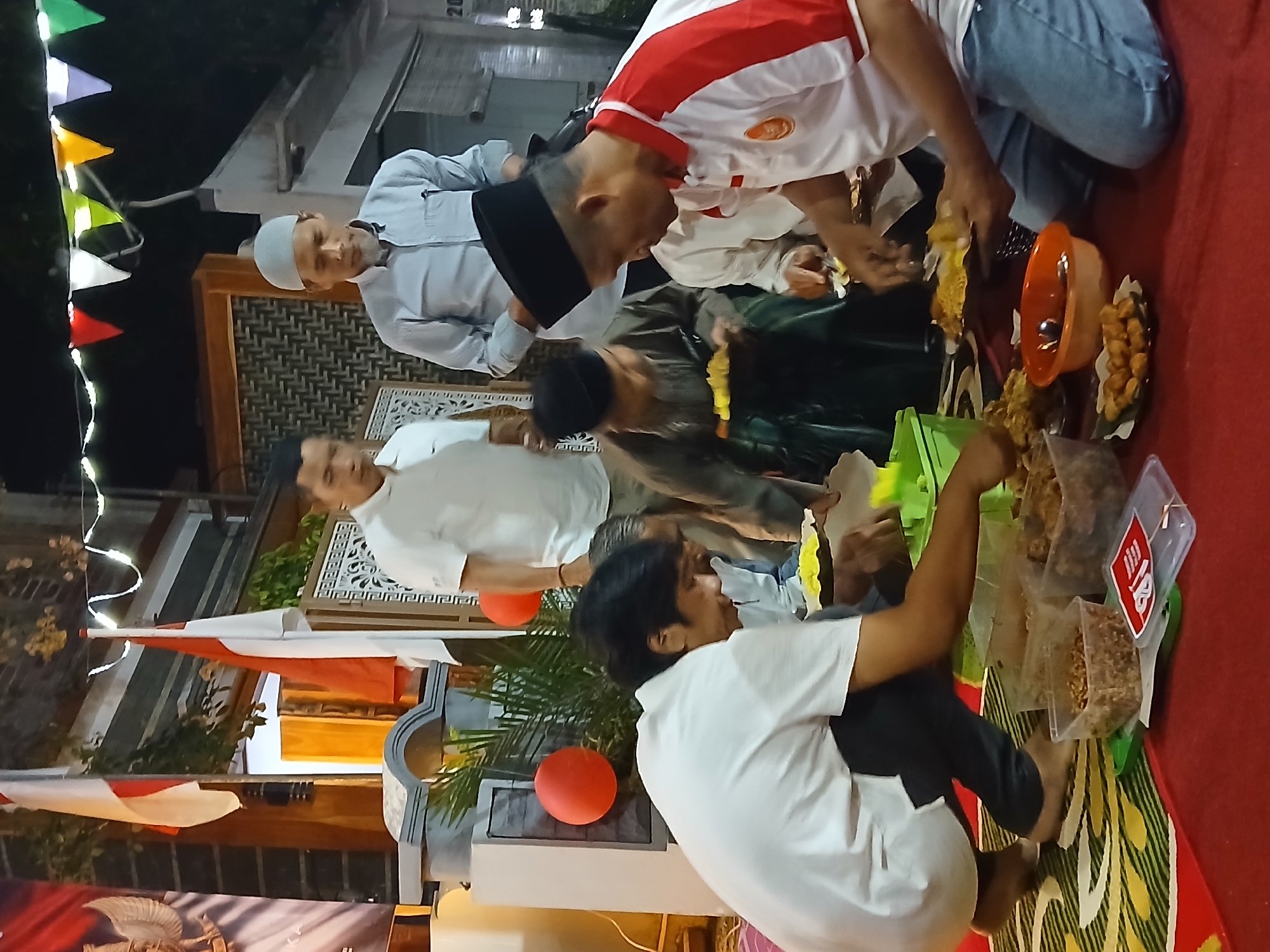 The image depicts an outdoor evening gathering in Ambarawa, Indonesia, with several men seated on the ground, engaged in eating. The scene is illuminated by string lights overhead and appears to be taking place on a decorated mat. Indonesian flags are visible in the background, along with a banner displaying an eagle emblem. Various food containers, plates of food, and bowls are arranged around the seated individuals. One man in the foreground is wearing a white polo shirt with red accents and jeans, while another person to his left is in a white t-shirt and black pants. Other men in the image are dressed in traditional attire, including caps and long-sleeved shirts. The ambiance suggests a communal meal or celebration.