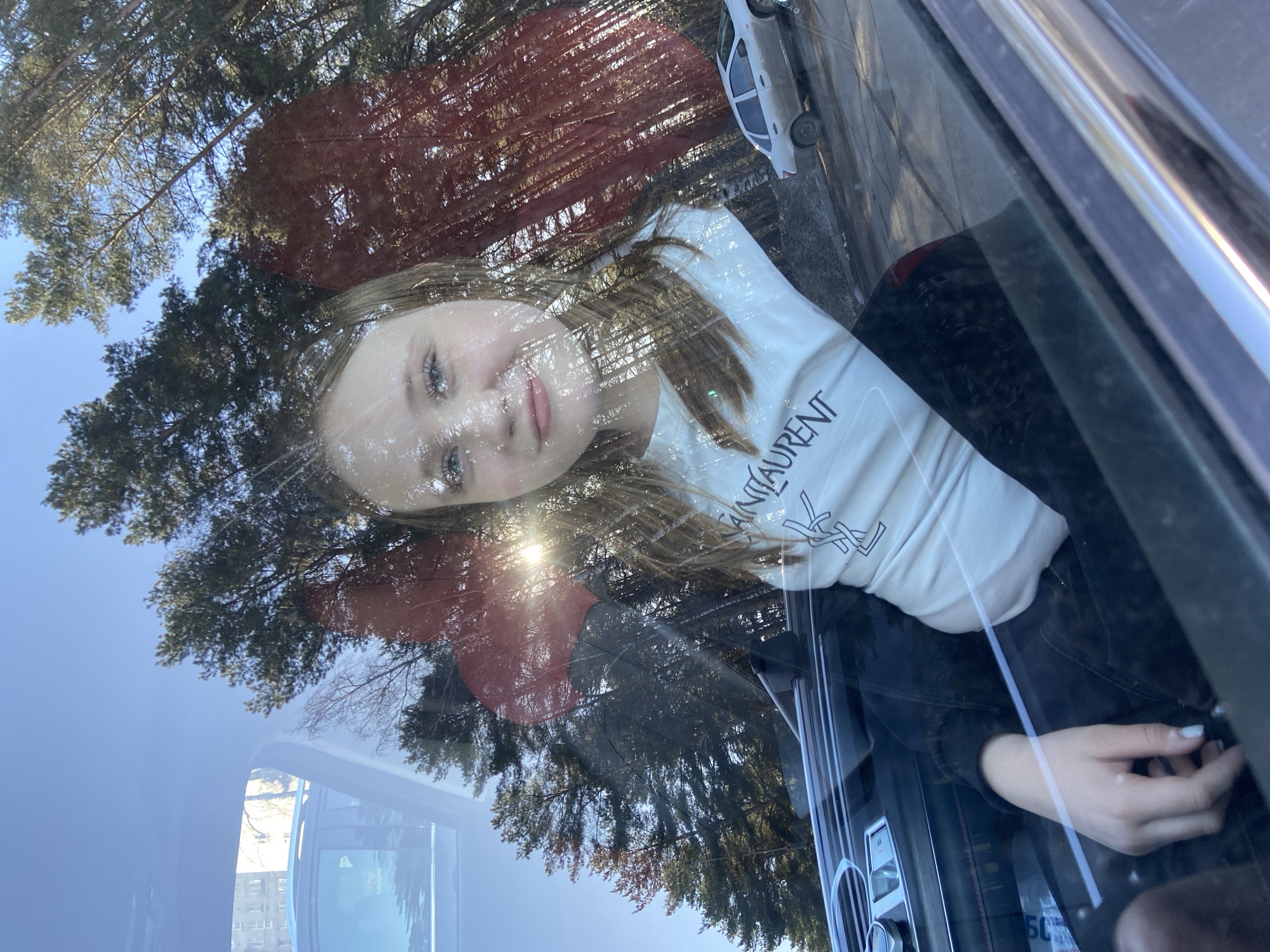 The image features a young female with light hair and fair skin, captured through a car window, giving her a translucent appearance due to strong reflections. She has light-colored eyes and a faint smile. She is wearing a light-colored long-sleeved top with the text "SAINT LAURENT" and the distinctive "YSL" logo prominently displayed on it. Her right hand, with light-colored fingernail polish, is visible inside the car.
The car's window reflects a bright, clear blue sky and a dense canopy of trees, including tall conifers like pines or firs, and some bare deciduous trees. Sunlight streams through the foliage, creating sun glare and illuminating the scene. In the upper right of the reflection, a small white car is visible, suggesting a street or parking area. The reflections are layered over the young woman and the car's interior, creating a complex visual effect.
The scene takes place during a sunny daytime, indicated by the clear sky and bright light. The natural elements in the reflection suggest a setting in a wooded or semi-rural area, consistent with the location context of Chaikovsky, Russia. The image combines an intimate portrait with an expansive outdoor reflection, blurring the lines between inside and outside.