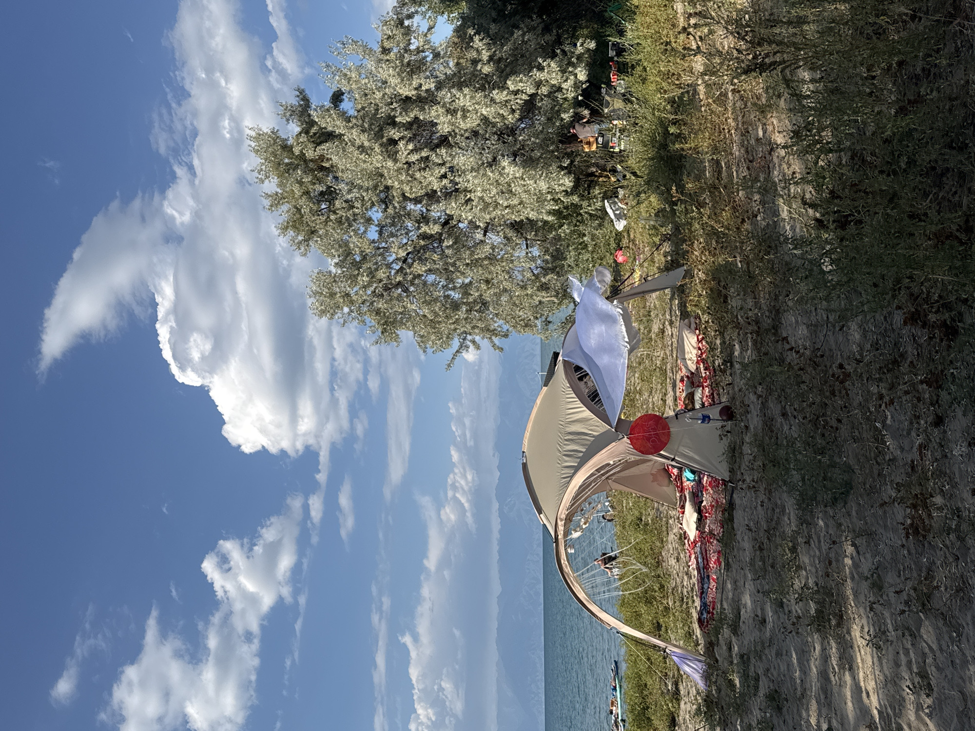 The image depicts a campsite on a sandy beach next to a body of water, likely Lake Issyk-Kul, near Basbeshik, Kyrgyzstan. The foreground is dominated by a tan and beige dome tent, partially open, with a red circular object hanging inside. A colorful blanket or rug is spread out in front of the tent, and a white sheet is draped over one of the tent poles, fluttering in the breeze. Various items, including pillows and bags, are visible around the tent.
In the middle ground, a large, silvery-green tree with dense foliage stands to the right of the tent. Beneath the tree, several people are visible, along with camping chairs and equipment. To the left of the tent, the body of water stretches towards a distant mountain range, visible under a blue sky with scattered white clouds. Several inflatable rafts or paddleboards are floating on the water in the distance, some with people on them. The overall impression is of a relaxed day at the beach, with a campsite set up for leisure.