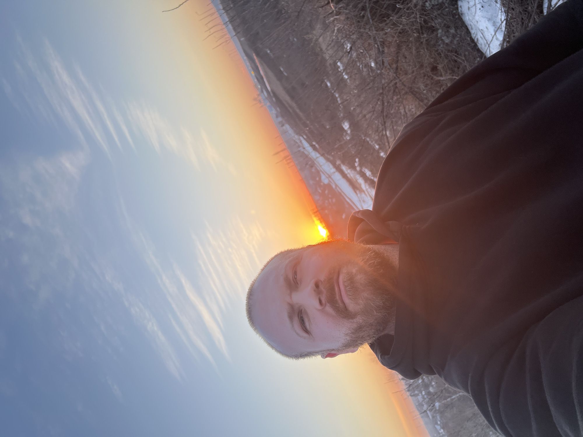 A man with short blond hair and a beard is posing for a selfie at sunset. The sun is visible on the horizon, casting a warm glow on his face. He is wearing a dark hooded sweatshirt. The background appears to be a winter scene with trees and snow. Without further information, it is impossible to verify the location as Chaikovsky, Russia.