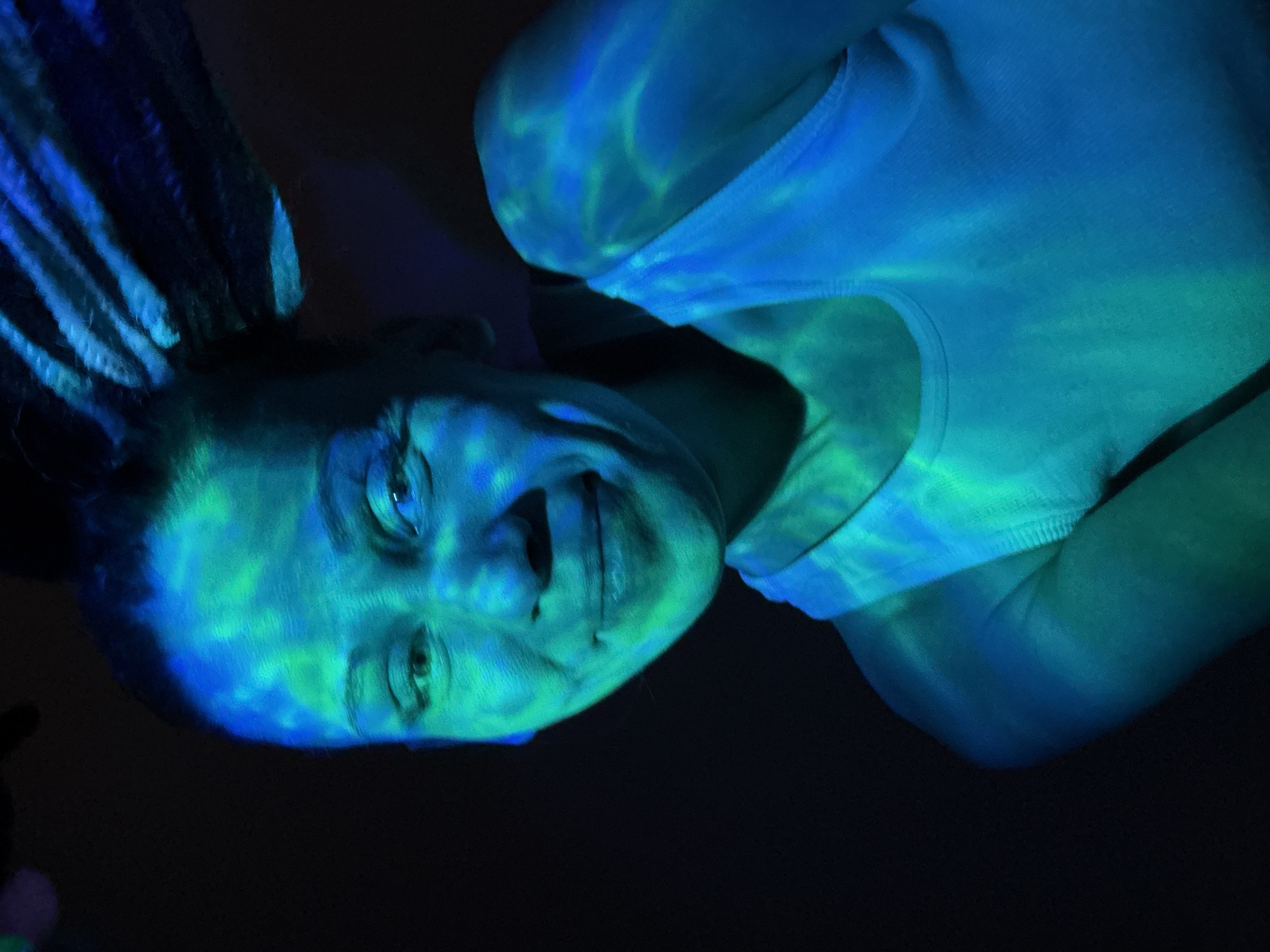 The image features a rotated close-up portrait of a person, likely female, set against a predominantly dark background. The subject's head is positioned towards the left of the frame, with her body extending to the right. Her face and upper torso are brightly illuminated by dynamic blue and green light, which casts wavy, almost water-like patterns across her skin, clothing, and hair.
The person is looking directly towards the viewer with a slight smile or smirk. Her eyes are visible, and her hair appears to be styled in dreadlocks or braids, also catching the vibrant light. She is wearing a light-colored or white tank top or undershirt, which is similarly covered in the blue and green light patterns. The dark background suggests an indoor setting with controlled, artificial lighting, possibly from a projector creating the patterned effect.
The overall atmosphere is artistic and somewhat ethereal, emphasizing the interplay of light and shadow. No specific activity beyond posing for a portrait is discernible. There are no visible objects other than the person and her attire. The time of day appears to be night or a darkened interior. No text is visible in the image. The location context provided is Bor, Russia, though this is not visually indicated within the image itself.