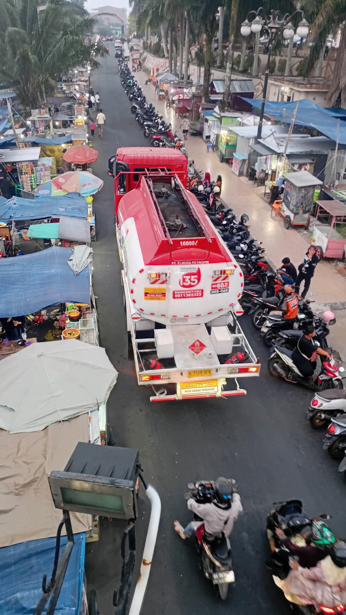 The image depicts a busy street scene in Jambi City, Indonesia, likely during daylight hours given the ambient light. The foreground features a large red and white tanker truck, presumably transporting fuel, parked on the right side of the road. The truck displays company logos and an emergency contact number. On the left side of the road, a market or roadside stalls are set up under tents and awnings, with goods displayed and people attending to them.
The road itself is lined with a long row of parked motorcycles, suggesting this is a popular area or a time of high activity. Several individuals are visible around the motorcycles and near the market stalls. Some are walking, others are seated or standing near their vehicles. On the far right, a few people are gathered, and some are on motorcycles. The overall impression is one of a vibrant, commercial area with significant foot and vehicle traffic, characteristic of a bustling Indonesian city. The environment appears to be an urban setting with trees lining one side of the street and buildings visible in the background.