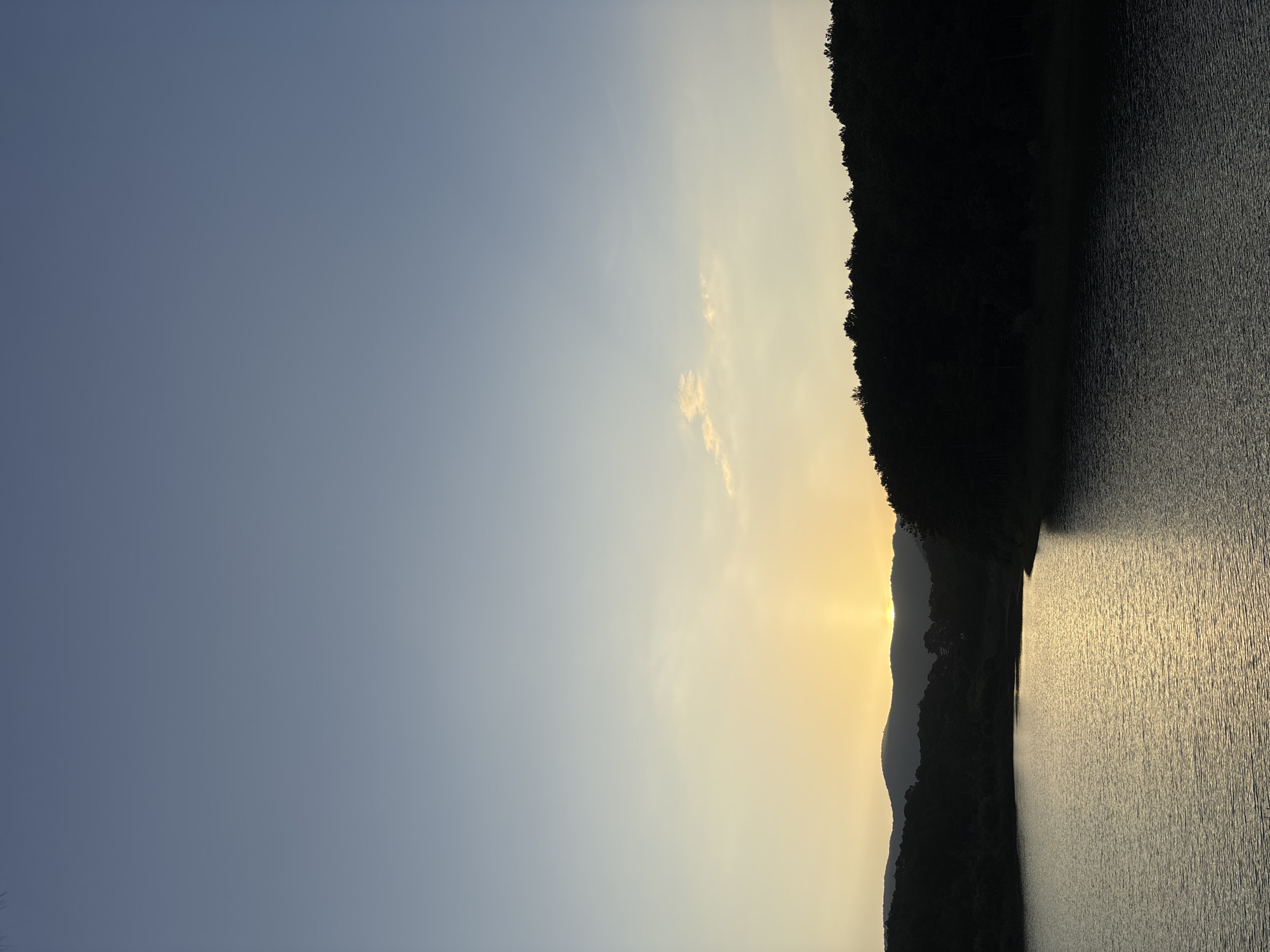 The image depicts a serene lake at sunset, framed by dark, silhouetted hills covered in trees. The sky transitions from a soft blue at the top to a warm yellow and orange near the horizon, where the sun is setting behind the hills. The water reflects the warm hues of the sunset, creating a shimmering surface. The scene is located at the Tuyen Lam Lake in Da Lat, Vietnam, a popular tourist destination known for its picturesque landscapes and natural beauty. There are no people or animals visible in the image, only the natural environment. The overall atmosphere is tranquil and peaceful, capturing the quiet beauty of a sunset over a lake.
