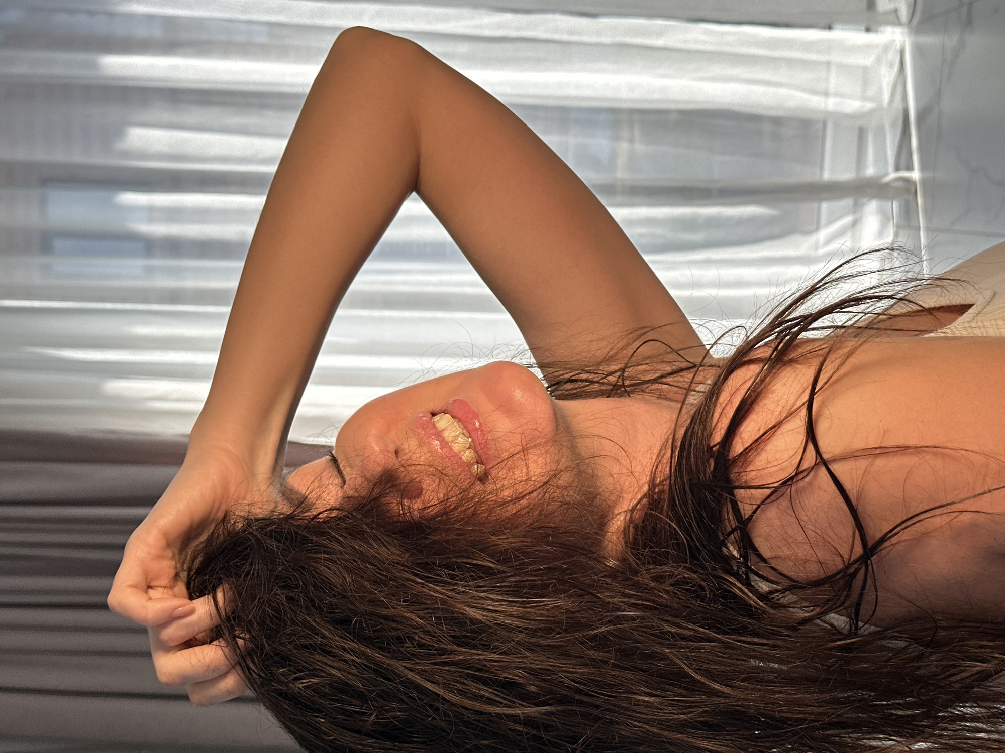 A close-up, horizontal shot depicts a person, likely a woman, lying down indoors in Chaikovsky, Russia. The subject's head is angled towards the bottom right, with her body extending upwards and to the left. She has long, dark, slightly disheveled hair that partially covers her face and is spread out around her head.
One arm is raised above her head, bent at the elbow, with her hand gently resting on her forehead or intertwined in her hair. Her skin has a warm, tanned complexion. A subtle smile is visible, showing her upper teeth and part of her lower lip. Her eyes appear closed or mostly obscured by her hair.
The background features a window covered by white horizontal blinds or curtains, allowing strong natural light to filter through and cast soft shadows, creating a pattern of light and dark horizontal stripes. This suggests the image was taken during the daytime. The overall impression is one of relaxation and contentment. No text is visible in the image.
