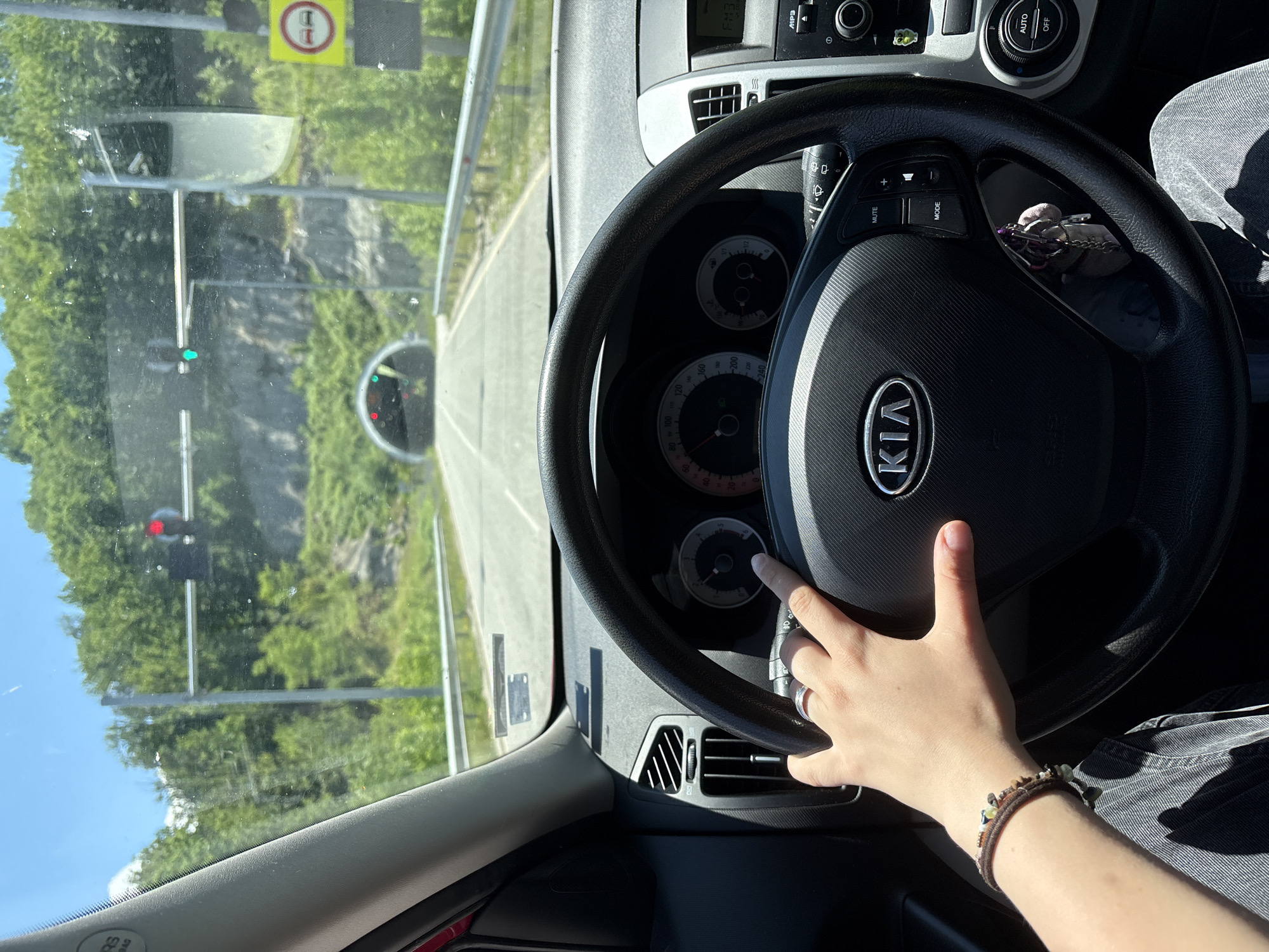 The image shows a first-person perspective from inside a Kia vehicle, likely a Kia Ceed, approaching a tunnel on a sunny day. The driver's left hand, adorned with a ring and a bracelet, is on the steering wheel. The dashboard features a speedometer and other instruments. Outside the car, the road leads to a tunnel entrance, with traffic lights visible overhead. A yellow sign with red circles and car icons indicates a no-passing zone. The surrounding environment is characterized by lush green trees and a rocky hillside, suggesting a mountainous or hilly terrain. The presence of a tunnel and traffic lights indicates this is a road infrastructure point, possibly on a highway or main route. While specific landmarks within Gabrovo are not directly identifiable, the general setting suggests a scenic route outside the urban center, potentially on the approach to or exit from the city, given the context of tunnels and natural surroundings often found on the outskirts of cities in such regions.