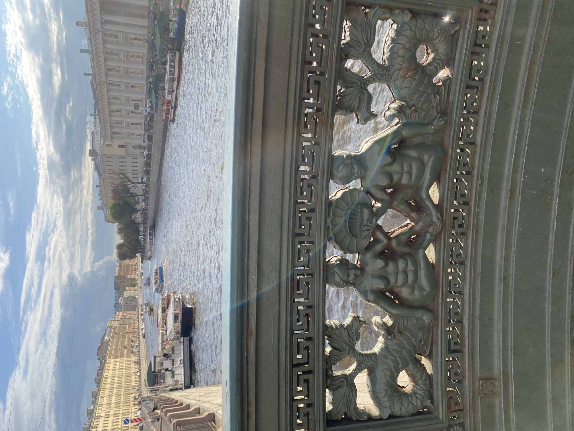 The image shows a view from a bridge over the Griboyedov Canal in Saint Petersburg, Russia. In the foreground, a dark green ornate metal railing features a bas-relief carving of two figures with fish-like tails, seated and holding a fan between them. A Greek key pattern adorns the railing above the figures. The canal flows through the city, with several tour boats navigating the water. Along the banks of the canal are multi-story buildings, typical of Saint Petersburg architecture, with yellow and white facades. The sky above is partly cloudy with the sun casting shadows and creating a subtle rainbow effect across the railing.