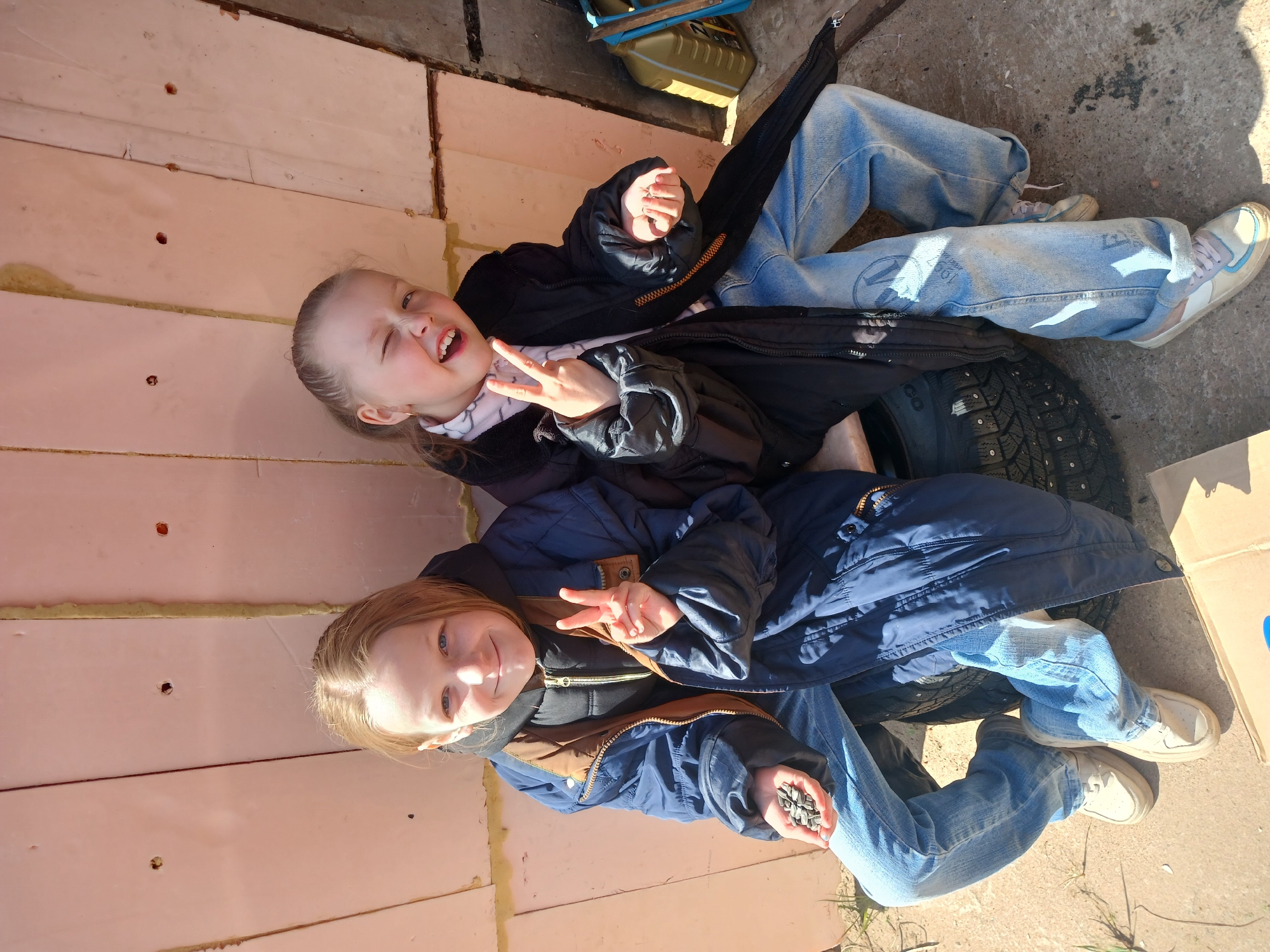Two young girls are sitting on a tire in what appears to be an outdoor setting. The first girl is on top wearing a black coat and light blue jeans, smiling and making a peace sign while winking. The second girl is seated below the first girl wearing a blue coat and light blue jeans, smiling and making a peace sign, while holding a handful of black seeds in her other hand. Both girls are wearing sneakers. The girls are located on a concrete or paved surface adjacent to a wall or structure made of pink insulation material, likely in an area undergoing construction or renovation. There is a cardboard box in the right corner of the image and a box in the top right corner. The presence of snow tires suggests a colder climate. The specific location of Noviy, Russia cannot be determined from the image.