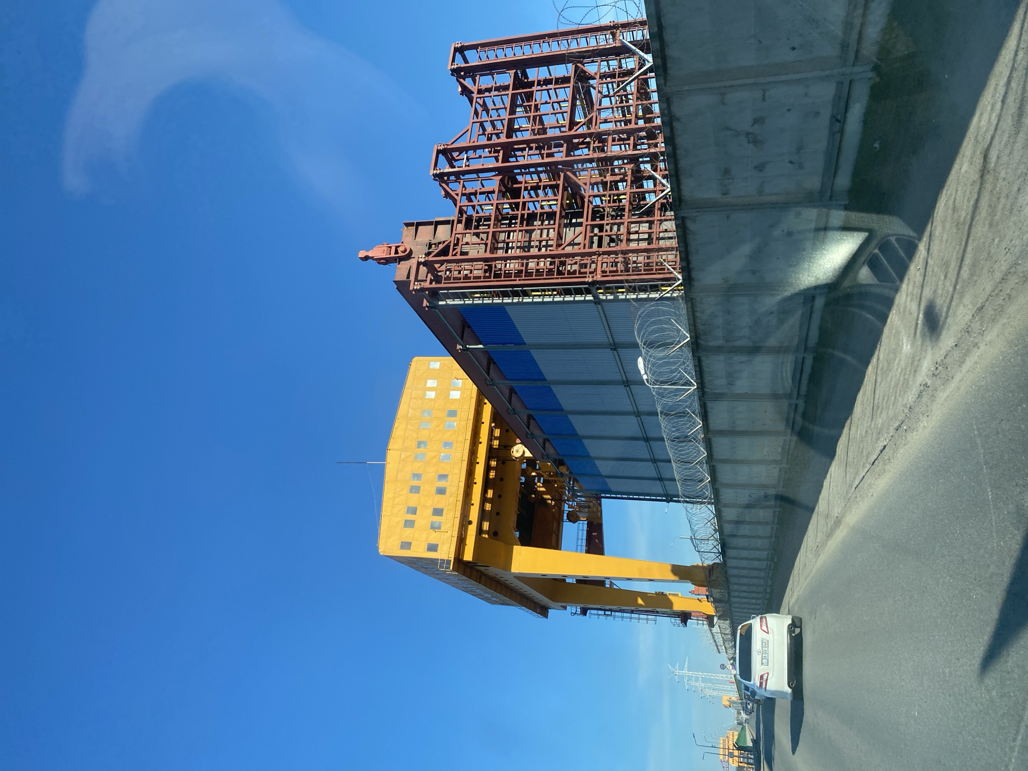 The image depicts a construction site in Chaikovsky, Russia, where a large, yellow, gantry crane is positioned next to a partially completed, red, steel bridge section. The bridge section is supported by concrete pillars and is enclosed with barbed wire. The sky is a clear, light blue. A white car is driving on a grey asphalt road alongside the construction site. In the distance, there are electrical power lines and street lights.