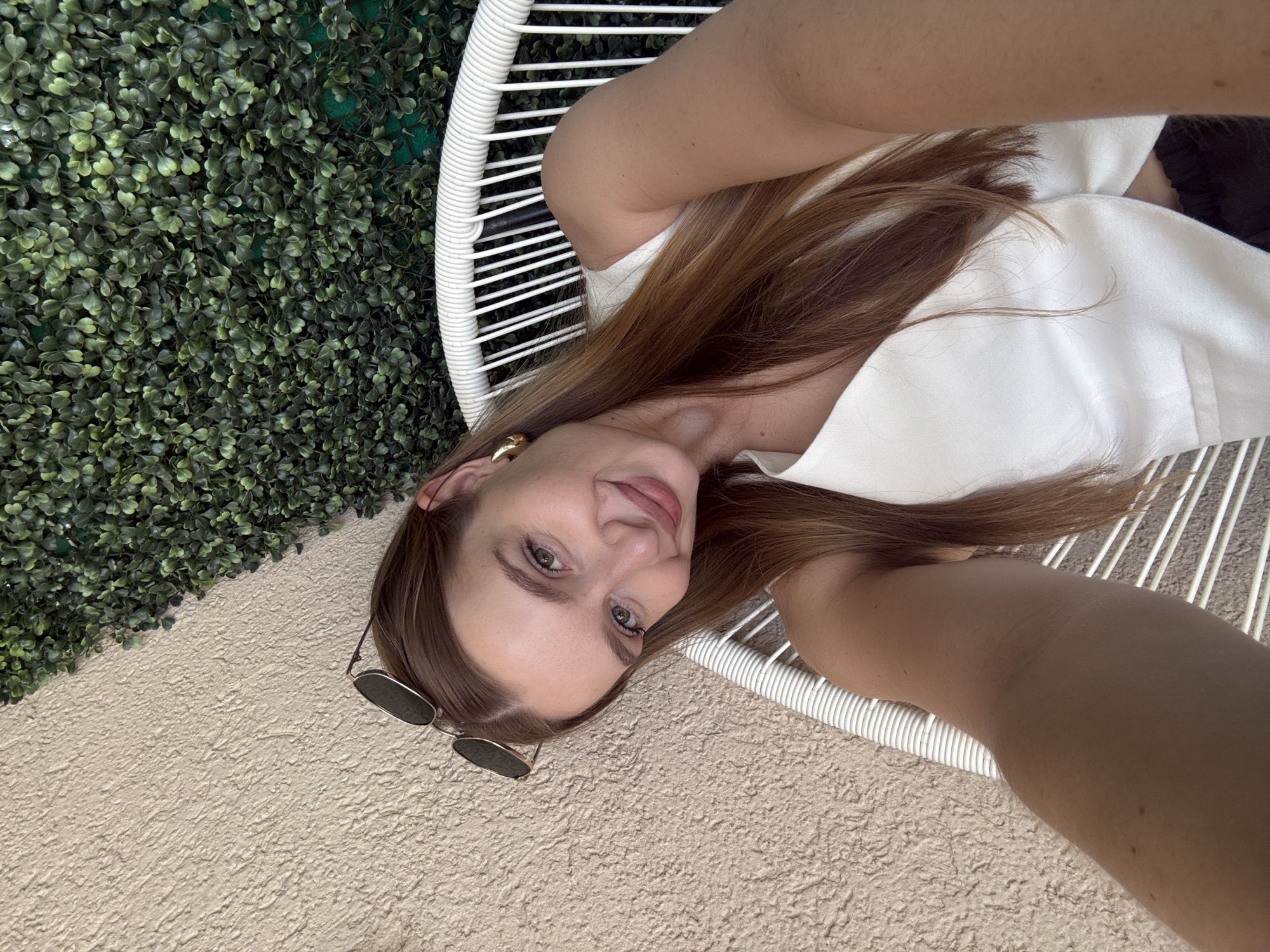A young woman is seated in a white wicker chair, appearing to take a selfie. She is fair-skinned, with light brown hair and green eyes. She is wearing a white top and a gold earring. Sunglasses are placed on the textured ground next to the chair. Behind her, there is an artificial green wall composed of small, leafy plants. The ground appears to be a light tan, textured surface, possibly concrete or stucco. Based on the presence of the artificial green wall and the overall aesthetic, the location may be in an outdoor seating area, possibly a cafe or bar. While it's difficult to pinpoint the exact location without more visual cues, the scene resembles an outdoor area in Budva, Montenegro.