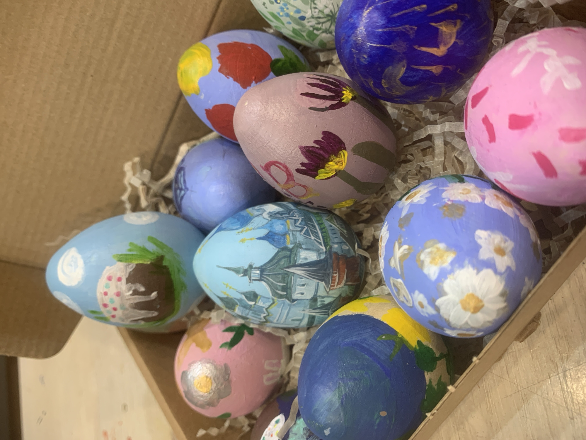 A close-up shot reveals a box filled with approximately a dozen painted Easter eggs nestled among shredded paper. The eggs display various designs, including floral patterns, abstract shapes, and what appears to be a cityscape resembling a Russian Orthodox church or cathedral. Given the presence of Russian architecture, the cityscape depicted on one of the eggs possibly represents a cathedral located in Izhevsk, Russia.