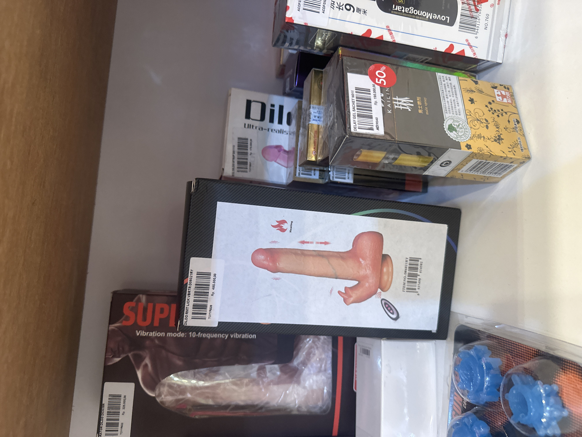 The image shows an assortment of adult toys and products arranged inside what appears to be a drawer. These items include vibrators, dildos with various features like a tongue (lick) and heating, and delay sprays. Based on the product packaging labels containing the abbreviation "Rp." and price listings such as "Rp. 325.000,00," "Rp. 495.000,00," and "Rp. 195.000,00" which are most likely in Indonesian Rupiah, the items appear to be located in North Kuta, Indonesia.
