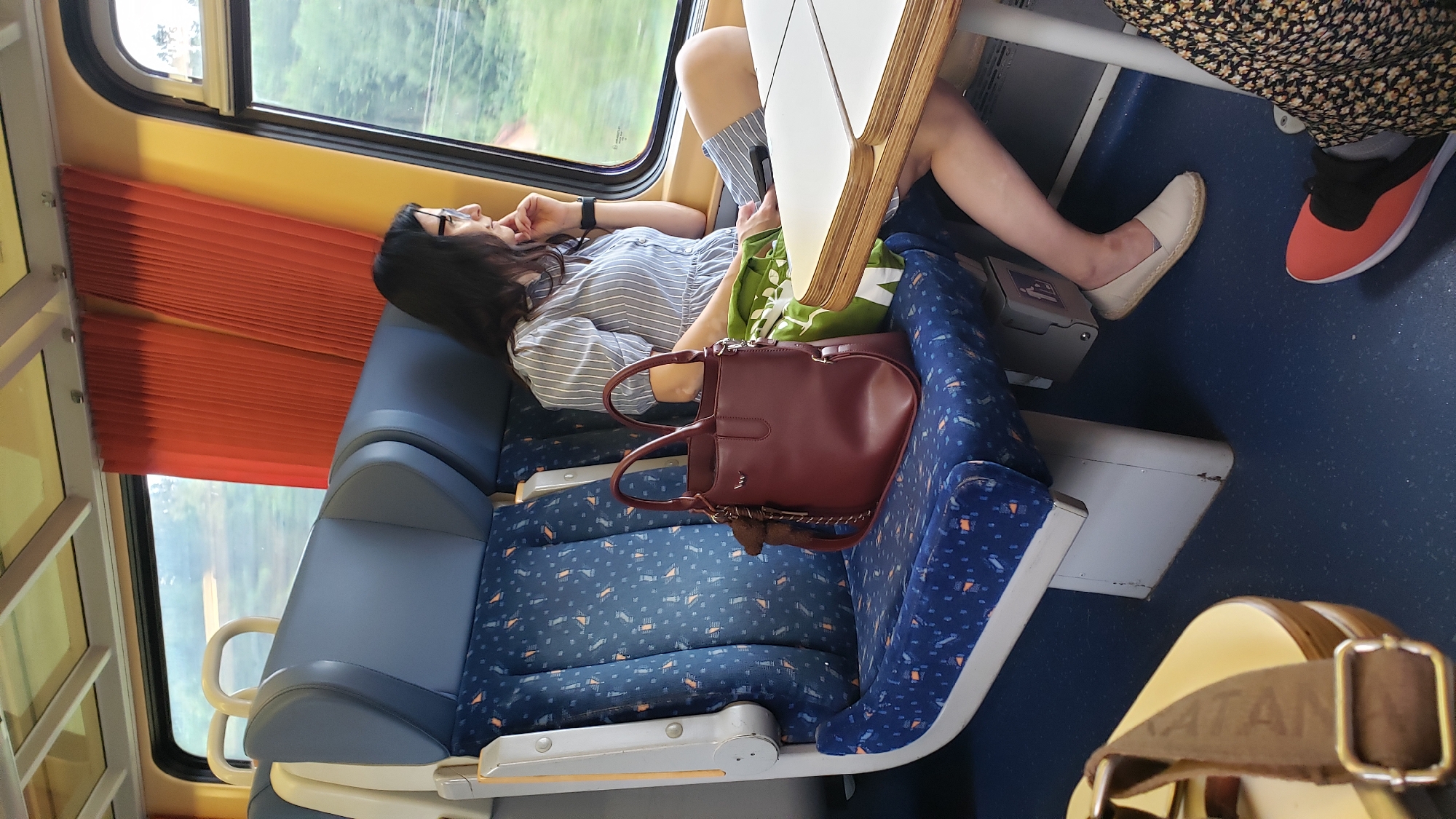 The image depicts the interior of a train compartment, likely in Nezbudská Lúčka, Slovakia. A woman with dark hair and glasses is seated by the window, looking outwards. She is wearing a striped dress and beige espadrilles. Next to her on the blue patterned seat is a burgundy handbag and a green shopping bag. A white table is positioned in front of her. In the foreground, another person's legs are visible, wearing a floral skirt and red sneakers. The train compartment features blue seats with a repeating pattern of small orange and blue shapes, and a bright yellow trim along the top of the windows. Orange pleated curtains are drawn back on the right side of the image. The overall impression is that of a passenger traveling by train through a green, tree-lined landscape.