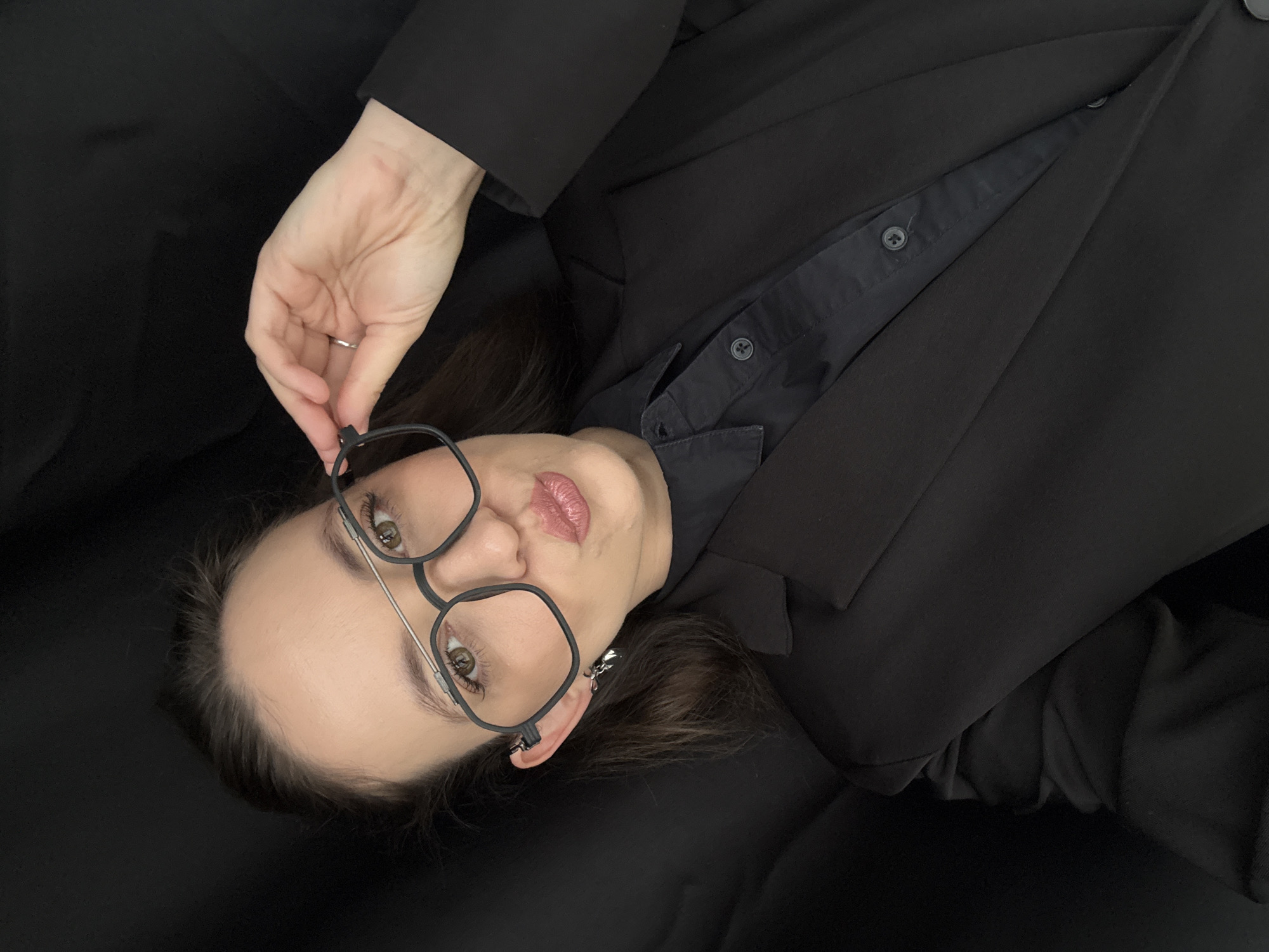 A woman is captured in a close-up, over-the-shoulder selfie, her head tilted slightly. She is wearing a black blazer over a black button-down shirt and large, square-framed glasses. Her left hand is raised, fingers delicately adjusting the temple of her glasses. She is wearing a small silver ring on her left ring finger and a dangling silver earring in her left ear. Her lips are pursed, and her brown eyes are looking towards the camera. The background is a dark, textured fabric, possibly a curtain or a draped cloth, creating a stark contrast with the woman's attire. The lighting is soft, highlighting her face and clothing. The exact location within the city of Chaikovsky, Russia cannot be determined from the visual information provided in the image.