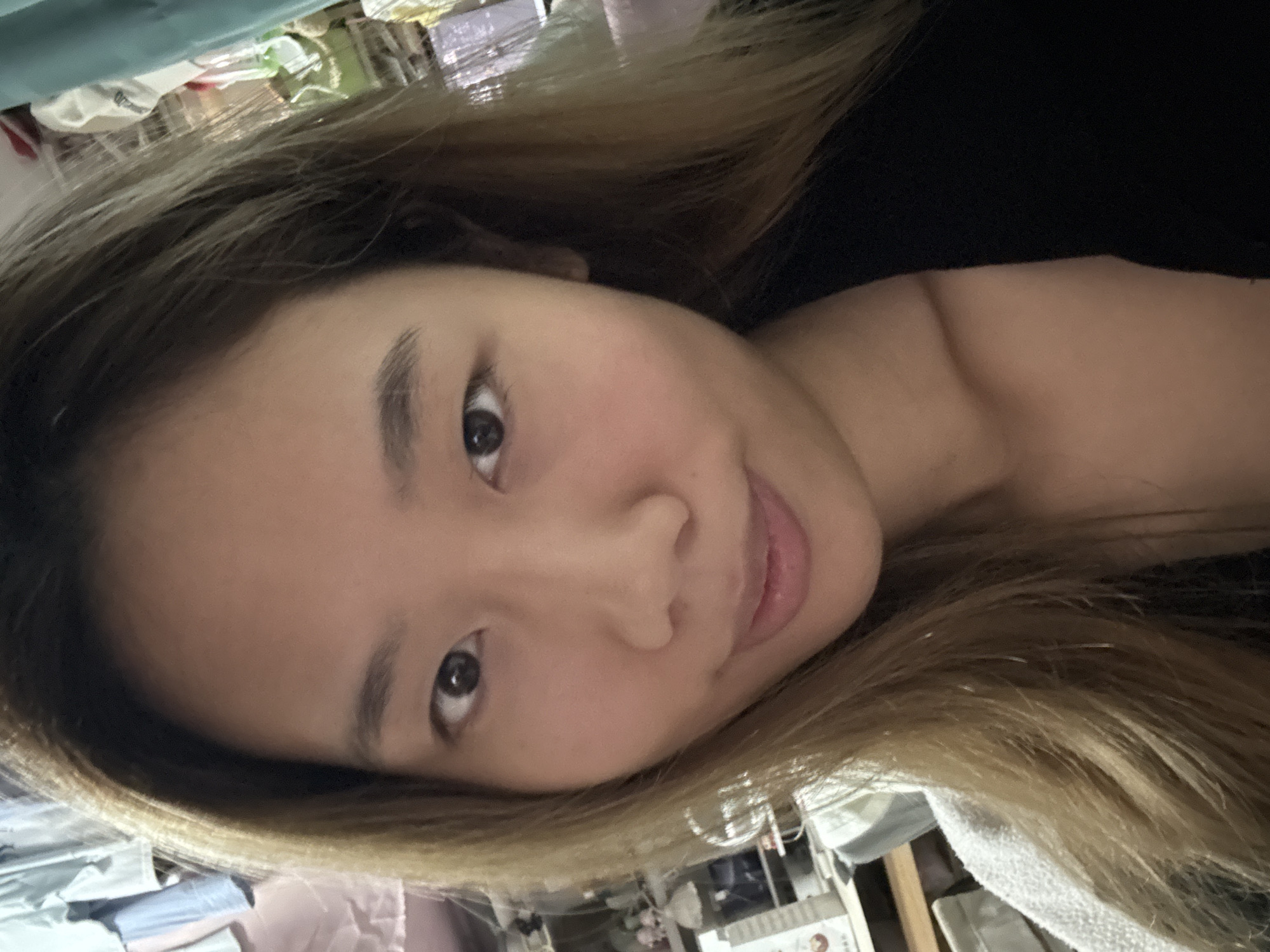 A close-up selfie of an Asian woman with brown hair is oriented at an angle. The woman is wearing a black top. The background includes some furniture, possibly a bed, and other objects. It's impossible to determine the location in Nan'an District, China, from the image alone.