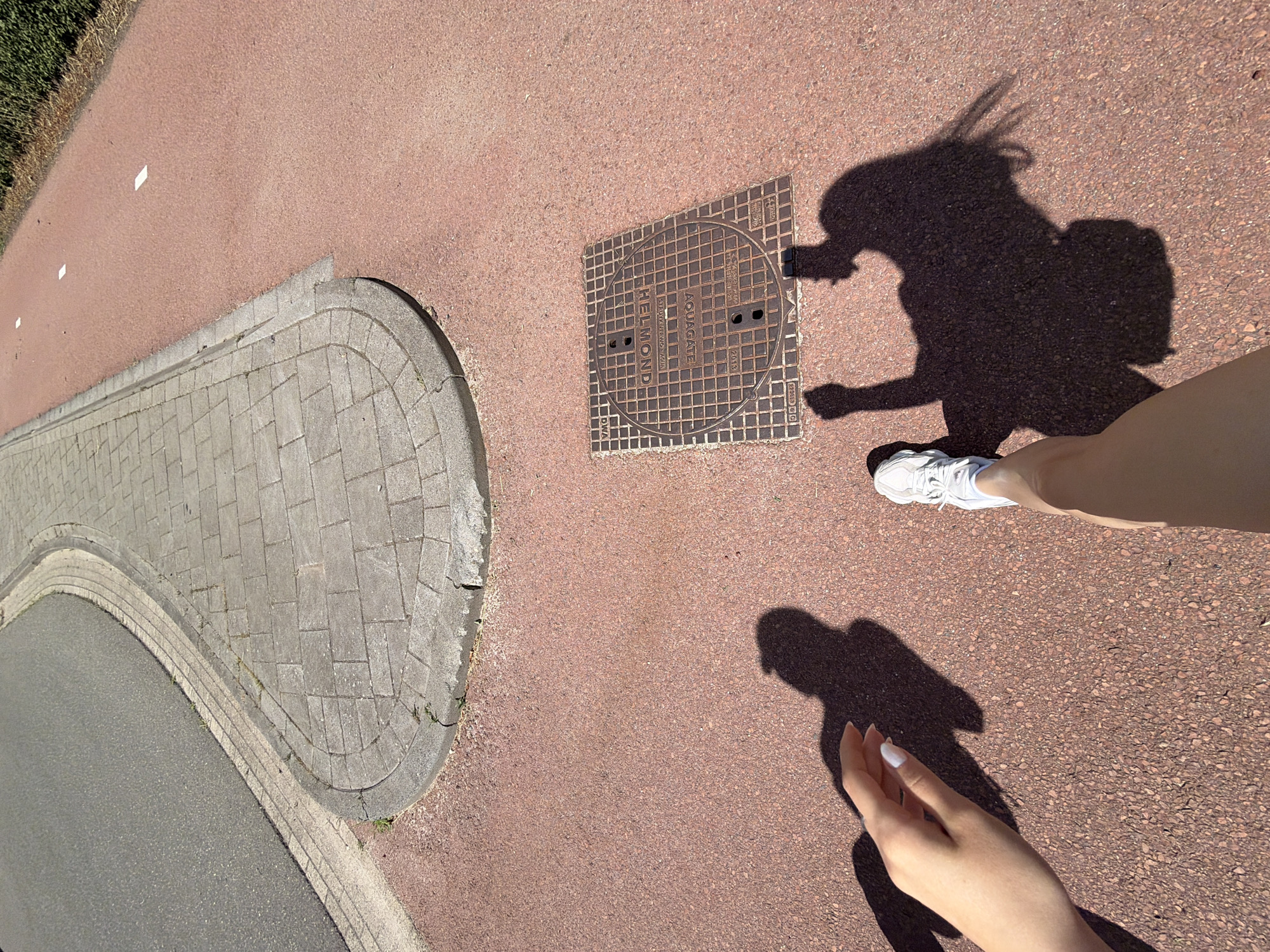 This media file is a low-angle, wide-shot photograph taken outdoors on a sunny day. The scene features a person's legs and feet, wearing white sneakers and a light-colored garment, casting a prominent shadow on a reddish-brown textured ground. Another shadow of a person with a backpack is visible, suggesting the photographer is taking a selfie. A manhole cover with "AQUAGATE" and "HELMOND" inscribed on it is present in the frame. A curved paved walkway and a road are also visible, indicating an urban or park setting. The lighting and shadows suggest it is daytime, likely late morning or afternoon, with bright sunlight. The overall mood is casual and observational.