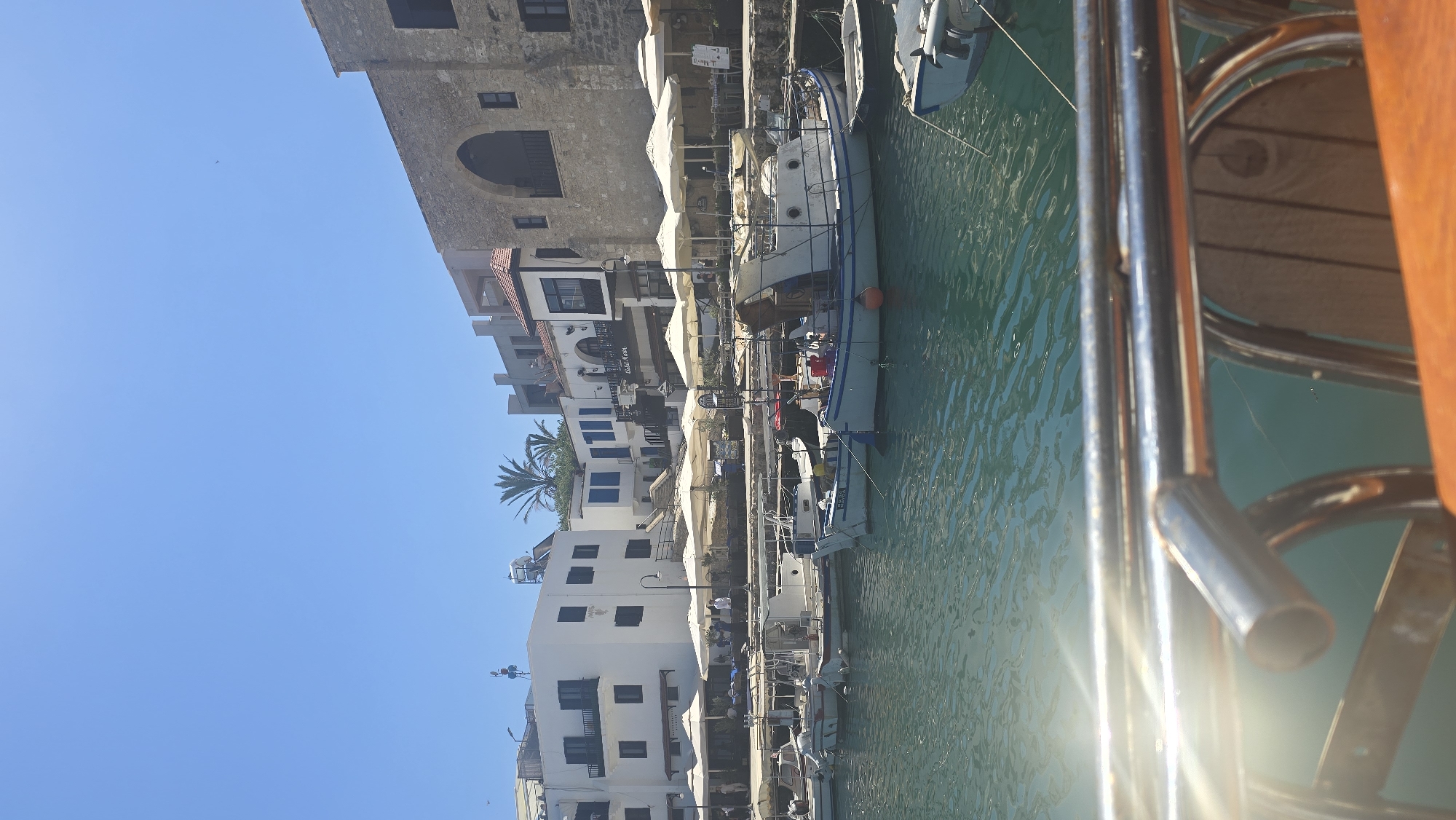 The image shows a bright, sunny day in the Kyrenia harbor in Northern Cyprus. Several small fishing boats are docked in the clear blue water. In the background, white buildings with blue shutters line the harbor, and a large stone building with an arched entrance stands to the right. Outdoor seating areas with white umbrellas are set up in front of the buildings. A wooden railing and part of a wooden chair are visible in the foreground, suggesting the photo was taken from a boat or a similar vantage point. The water is slightly rippled, and sunlight reflects off its surface. A palm tree is visible behind some of the white buildings. The sky is a clear, vibrant blue.