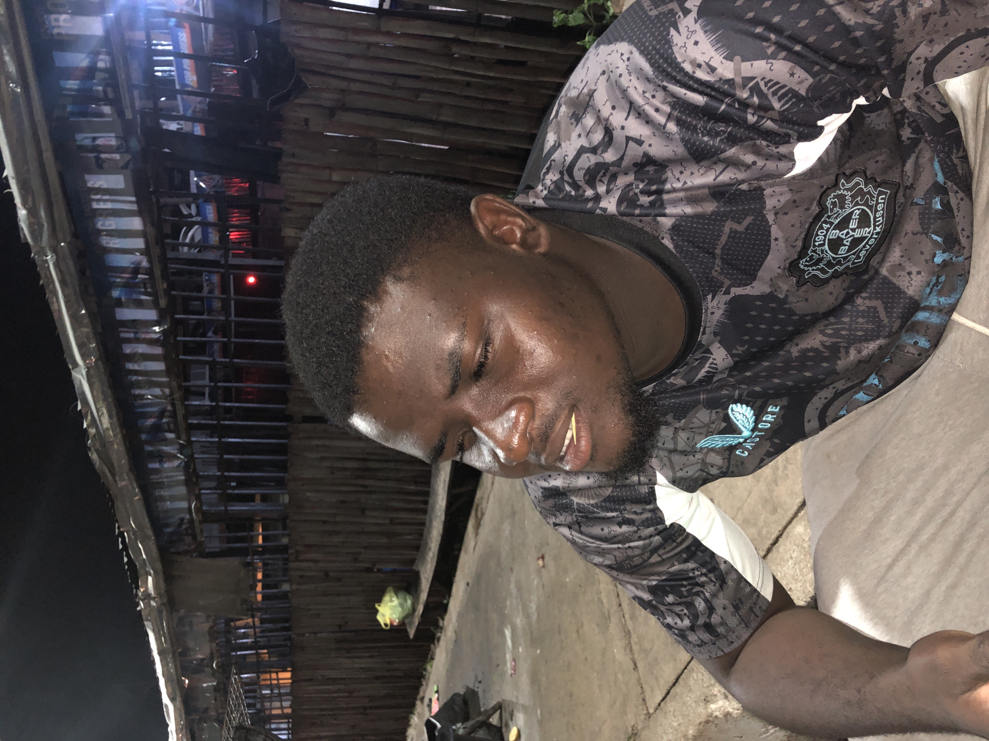 The image shows a young Black man with short, dark hair and a beard, wearing a grey and black patterned shirt with the "Bayer Leverkusen" logo visible on the chest. He has a toothpick in his mouth and his eyes are closed, with sweat visible on his forehead and face, suggesting it might be warm or humid. The man is leaning forward, appearing relaxed or possibly tired.
The background is somewhat blurred but appears to be an outdoor setting at night. There are wooden structures behind the man, possibly part of a building or shelter. In the distance, there are lights and what looks like shelves filled with items, possibly in a market or shop. A yellow plastic bag is hanging from a hook to the left of the man. The ground is paved.
The scene is dark, indicating it is nighttime, and the lighting seems to come from artificial sources. The overall impression is a candid shot taken in a casual outdoor environment in Jos, Nigeria. The man's expression and the presence of sweat suggest a warm climate and potentially a relaxed or contemplative mood.