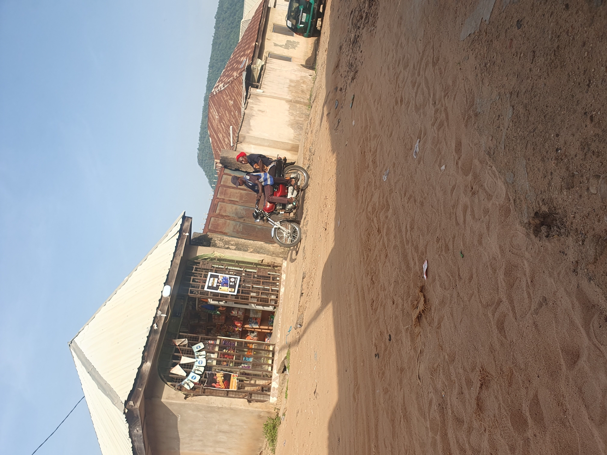 Two people are on a red motorcycle on a dirt road. The person in front is wearing a red cap and a dark blue shirt. The person behind is wearing a blue and white striped shirt and a dark shirt. They appear to be talking. The motorcycle is positioned facing towards the right side of the image. Behind them, there is a building with a corrugated metal roof and a concrete structure. The building appears to be a shop with shelves of goods visible through a metal grate. There are posters on the wall and on the front of the shop. One poster shows a man in a blue jersey. Another poster shows a man's face. In the background, there is a green vehicle parked in front of a building. The scene is set in an outdoor environment with a clear sky. The ground is covered in sand and there are some footprints visible. A steep, green, tree-covered hillside is visible in the far background. The image appears to be taken during the day, likely in the late morning or early afternoon, given the bright sunlight casting shadows. The setting suggests a marketplace or a residential area with small businesses in Lokoja, Nigeria.