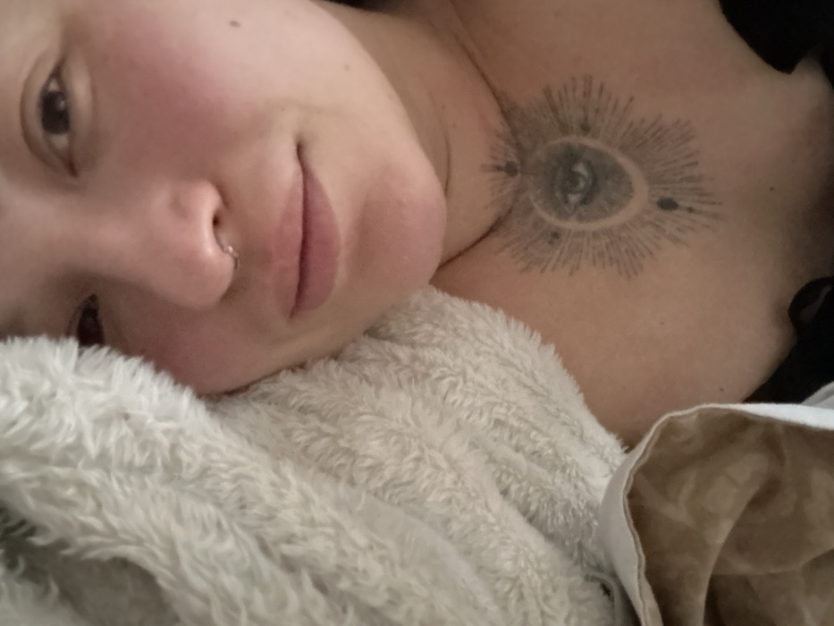 The image displays a close-up portrait of an individual, likely female, from the upper chest upwards. The person has fair skin, with visible flush on the cheeks, dark eyes, and full lips. A silver ring-style septum piercing is present. A prominent tattoo is visible on the upper chest/lower neck area, depicting a stylized eye within a crescent shape, surrounded by radial lines resembling a sunburst or starburst, with small circular accents. The left side of the individual's face and shoulder are partially covered by a thick, fluffy, light-colored material, consistent with a blanket or soft garment. Below this, a section of light brown, possibly patterned, fabric is visible. A dark garment, likely black, is visible at the lower right edge of the frame. The person is looking directly towards the viewer with a neutral to slightly smiling expression, appearing to be in a state of rest or relaxation. No discernible environmental details are present to identify a specific location within the city of Chaikovsky, Russia, or any other geographic area. The scene appears to be indoors.