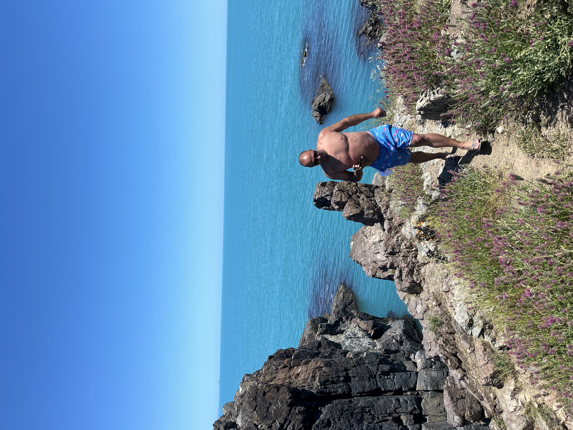 A shirtless, bald man wearing blue patterned swim trunks and sunglasses is walking on a dirt path along a rocky coastline. He is holding a smartphone in his right hand. The path is bordered by green vegetation and purple wildflowers. In the background, the bright turquoise sea stretches to the horizon under a clear blue sky. The scene appears to be the coast near Sozopol, Bulgaria.