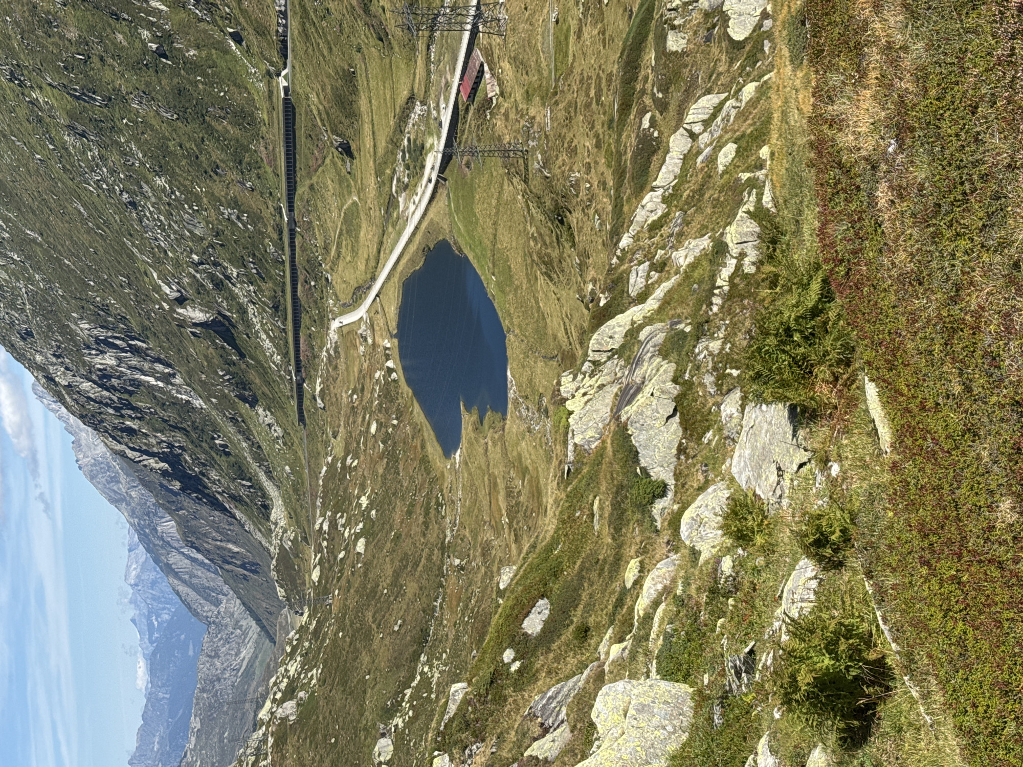 An aerial, wide shot captures a panoramic view of a mountainous landscape near Airolo, Switzerland. The scene is dominated by rugged, green slopes dotted with large, light-colored rocks and patches of dense vegetation. In the mid-ground, a dark blue, heart-shaped lake is nestled within the valley. A paved road, appearing as a white ribbon, encircles the lake and winds through the terrain. Along the road, a long, dark structure, possibly a tunnel entrance or gallery, is visible on the right side, with a small red-roofed building and power lines nearby. The distant background reveals a range of majestic, hazy mountains under a bright blue sky with scattered clouds. The overall impression is one of a serene, natural environment with human infrastructure integrated into the landscape.