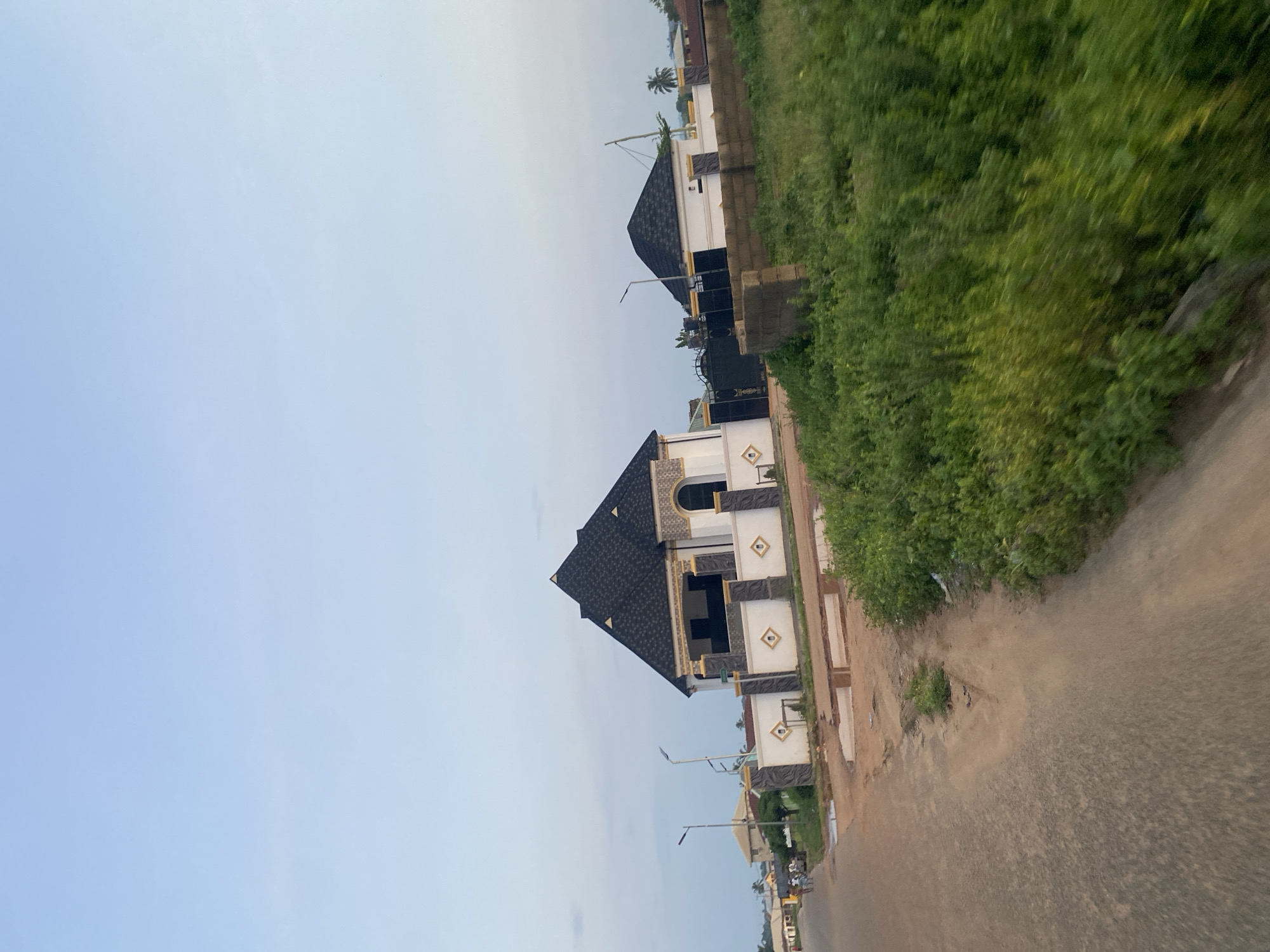 The image displays a residential street scene in Enwan, Nigeria. The foreground and left midground feature a two-lane road, characterized by a dark, textured asphalt-like surface on the left and a lighter, reddish-brown dirt or gravel path on the right edge, with visible puddles near a low curb.
On the right side of the road, two distinct houses stand behind perimeter fences. The prominent house on the left features a multi-faceted black shingle roof, white exterior walls adorned with gold and dark grey geometric patterns (including diamonds), and an arched window. Its perimeter fence matches the house's decorative style, incorporating columns and patterned panels. To its right, a smaller house is visible, also topped with a black shingle roof, partially obscured by a section of unfinished concrete block wall and a large patch of dense, vibrant green foliage that occupies the immediate foreground. A large black metal gate is situated between the two houses, providing access to one of the properties.
Along the left side of the road, at least two modern streetlights with solar panels are erected. Further down the road, a small group of approximately three to four indistinct individuals can be seen walking. The sky above is a clear, light blue with minimal cloud cover. The perspective suggests the photograph was captured from a moving vehicle, indicated by slight motion blur in the foreground vegetation.