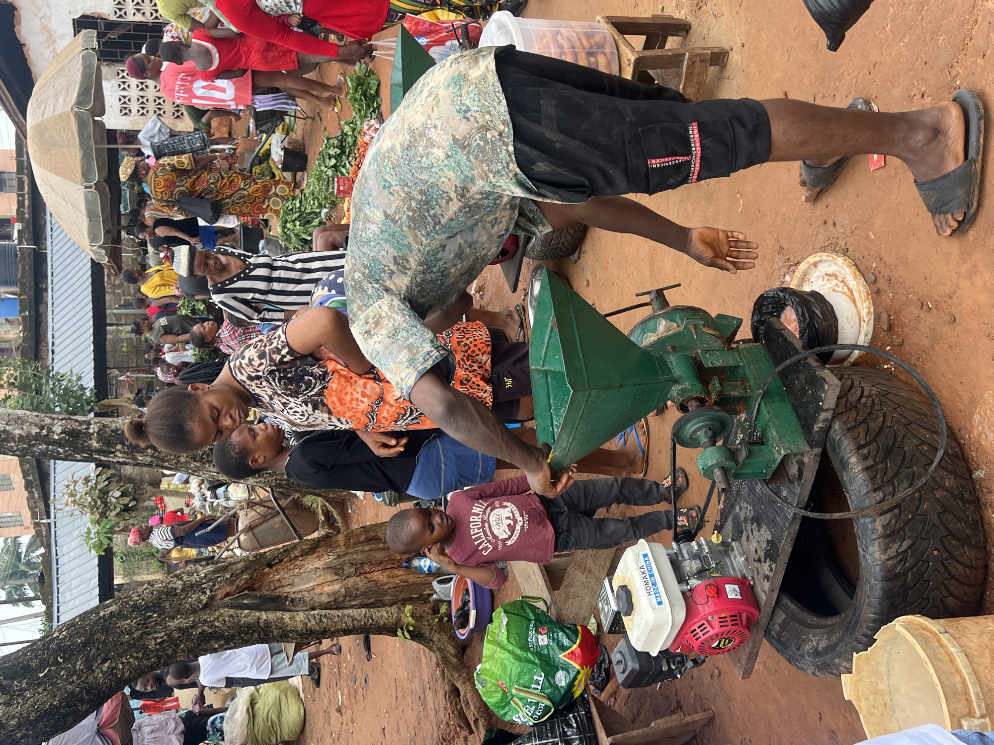 This image depicts a bustling outdoor market scene in Nkpor, Nigeria. The ground is a reddish-brown dirt. In the foreground, a man in a camouflage shirt and black shorts is operating a green grinding machine. The machine is powered by a red and white engine labeled "HOMAKA MADE IN CHINA," which is mounted on a wooden platform atop a large tire. To the left of the grinding machine is a large green plastic bag. A young boy in a maroon shirt and dark pants stands nearby, looking towards the machine.
Behind them, a woman in a patterned dress appears to be interacting with the boy. Various market stalls and a diverse crowd of people are visible in the background. Some stalls have awnings and display goods, including piles of green vegetables. Other market-goers are seen walking, standing, and observing. The overall atmosphere suggests a lively commercial transaction environment.