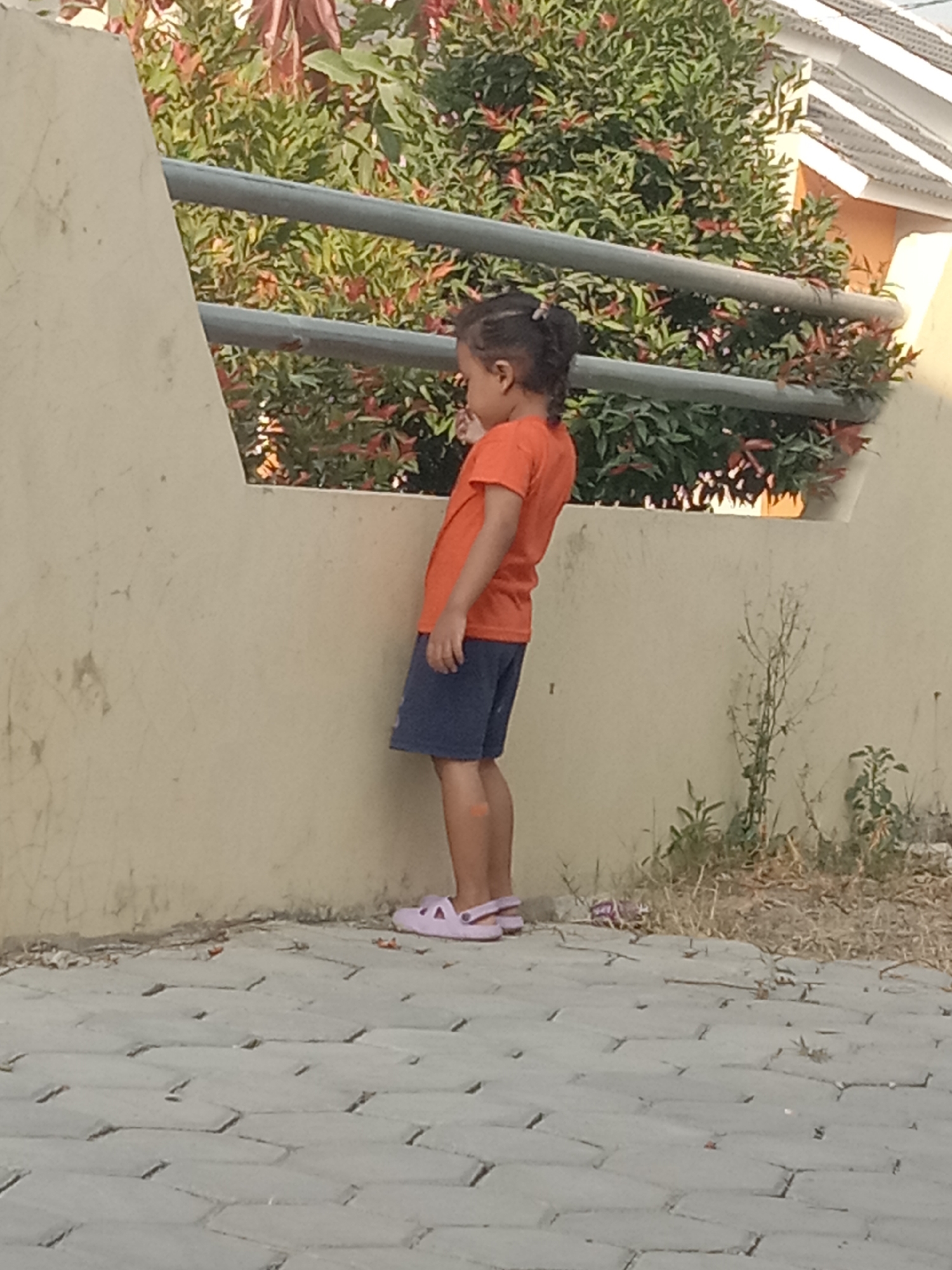 A young girl stands with her back to the camera, facing a textured, light-brown wall. She wears an orange t-shirt and dark blue shorts, along with light purple slip-on sandals. Her dark hair is tied back in a ponytail. The wall has a diagonal section extending upwards, with a horizontal metal railing running along its top. Behind the railing, dense green foliage with some reddish leaves is visible, and beyond that, the roofline of a building. The ground in front of the wall is paved with hexagonal interlocking stones, and to the right of the girl, dry grass and small plants grow near the base of the wall. The scene appears to be outdoors, possibly in a residential area of Sukaraya, Indonesia, suggested by the style of the house roof and the vegetation.