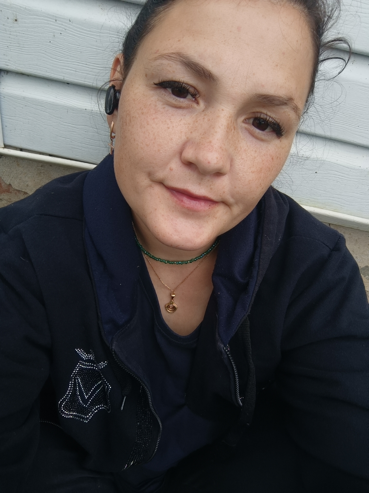A close-up, waist-up selfie of a fair-skinned woman with dark hair pulled back, wearing a dark blue hooded jacket over a dark blue t-shirt. She has freckles across her face, wears prominent false eyelashes, and has a black earbud in her right ear. A thin green beaded necklace with a gold pendant is visible. The background is a light-colored, horizontally ribbed surface, possibly siding, with a darker, rougher surface below it. The image has a slightly overcast, outdoor lighting. There is no discernible indication of the city of Karsha, Russia, or any specific urban environment.