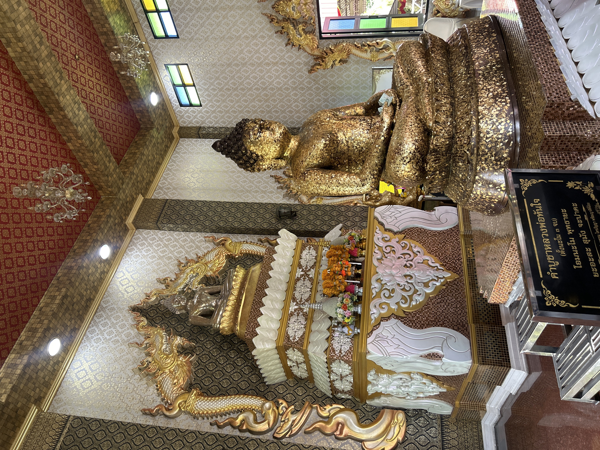 The image shows the interior of a Buddhist temple in Thai Ko Subdistrict, Thailand. Two large golden Buddha statues are the focal point. The statue on the right is seated in a meditative pose, covered in gold leaf, and appears to be the primary devotional object. To its left, a smaller, silver-colored Buddha statue sits on a multi-tiered, ornate pedestal adorned with carvings and flowers. The temple's interior is richly decorated with gold accents, patterned wallpaper, and red and gold patterned ceilings with chandeliers. Stained-glass windows are visible in the background, letting in colorful light. A black plaque with Thai writing is situated in the foreground, in front of a tiered platform and metal steps. The overall atmosphere is one of reverence and ornate religious artistry.