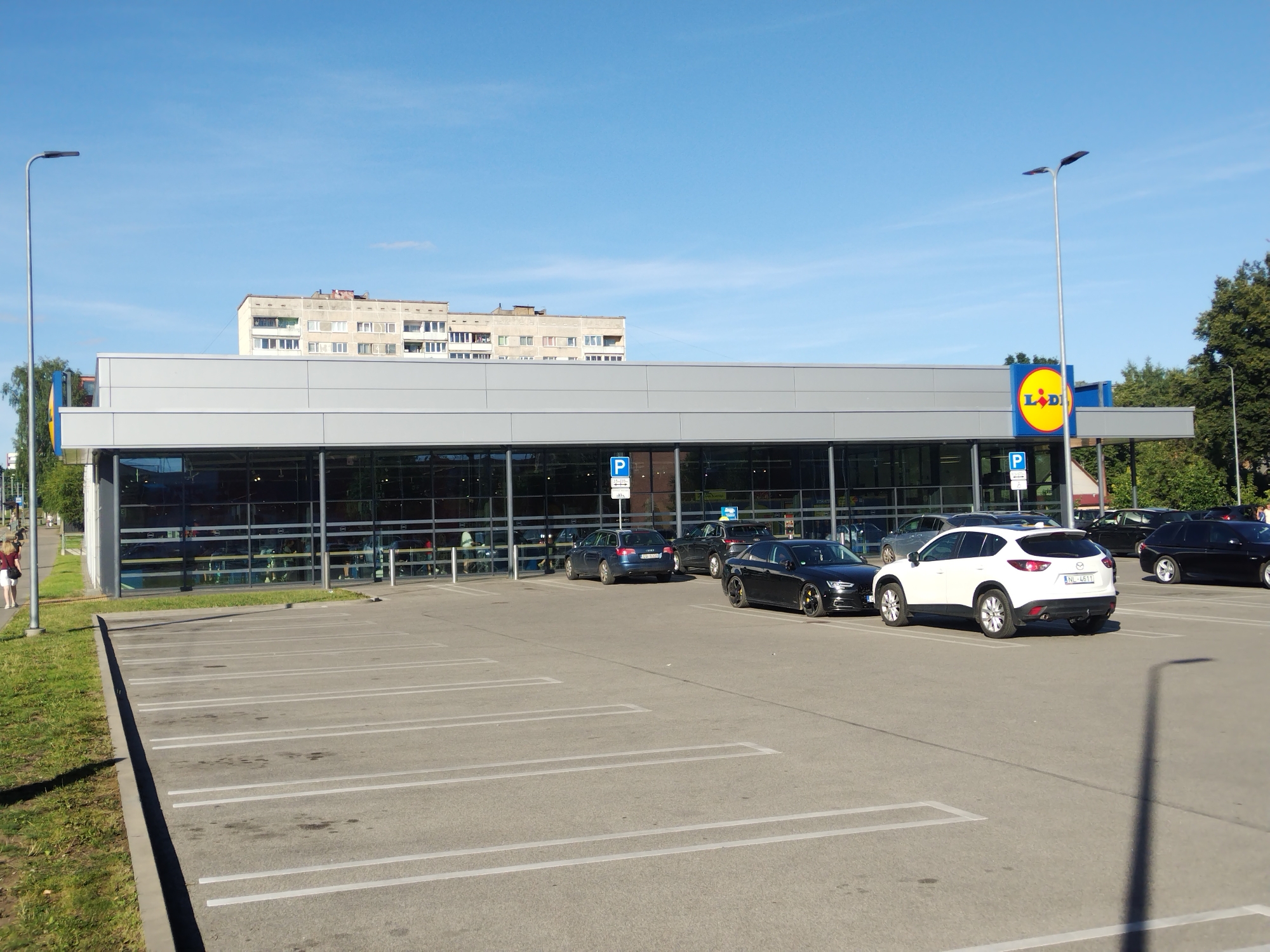 The image depicts a sunny day in the city of Daugavpils, Latvia, with a view of a Lidl supermarket. The building features a modern design with a grey facade and large glass windows. A prominent Lidl logo is visible on the right side of the building. In front of the supermarket, a parking lot is laid out with marked parking spaces. Several cars are parked in the lot, including a white Mazda CX-5, a black Audi A4, and a dark blue Audi A6. There are also two streetlights present, one on the left side and another on the right, indicating the presence of public lighting. In the background, a multi-story residential building stands, typical of Soviet-era architecture. A woman in a white top and red shorts is walking on the sidewalk to the left of the supermarket. The sky is clear and blue, with a few wispy clouds. The overall scene conveys a sense of everyday life in a suburban area.