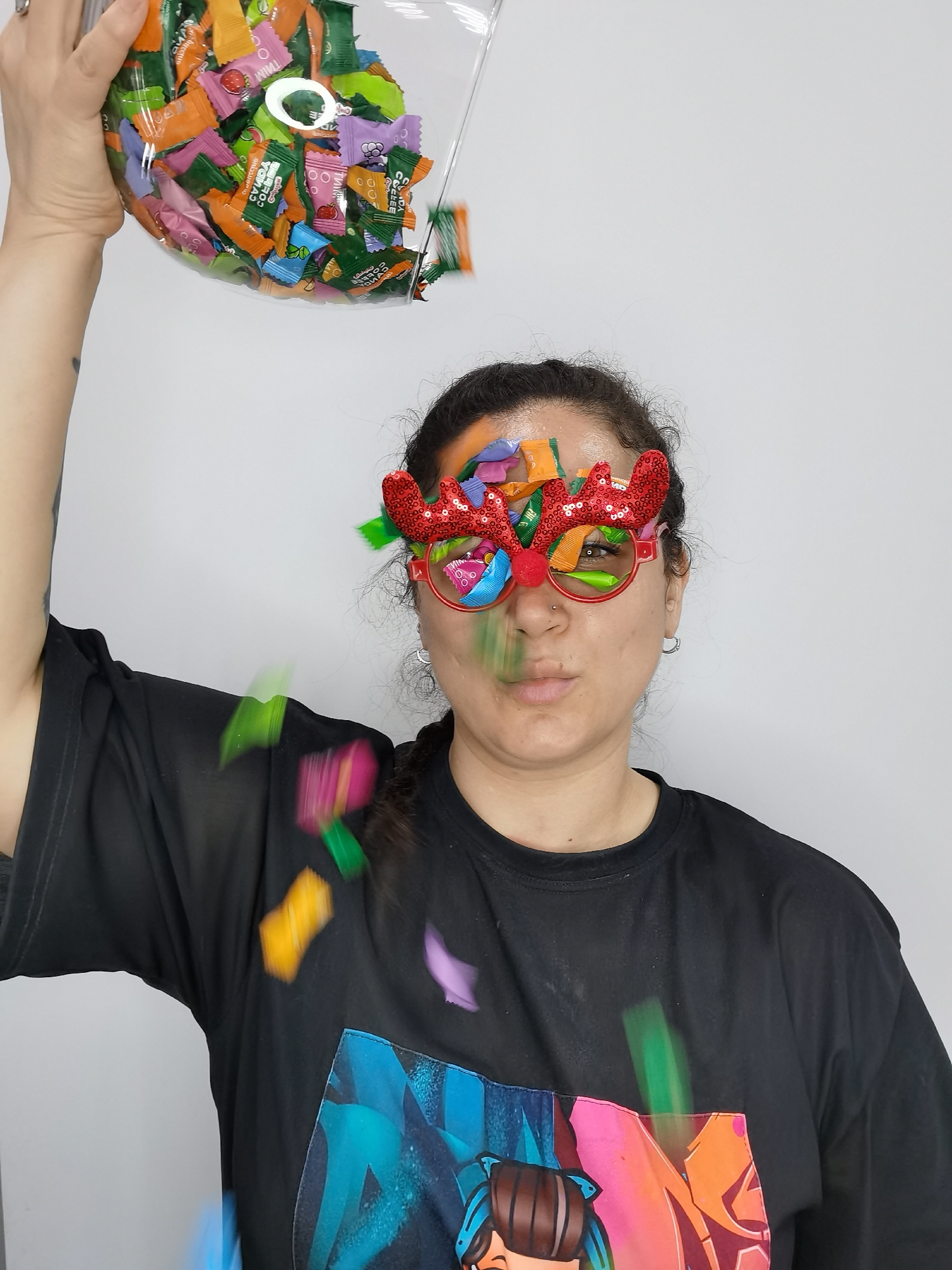 A person wearing reindeer glasses is having candy poured on their head from a clear plastic container. The person is wearing a black t-shirt with a colorful design on it. The candies are various colors and wrapped in individual wrappers. The background is a plain white wall.