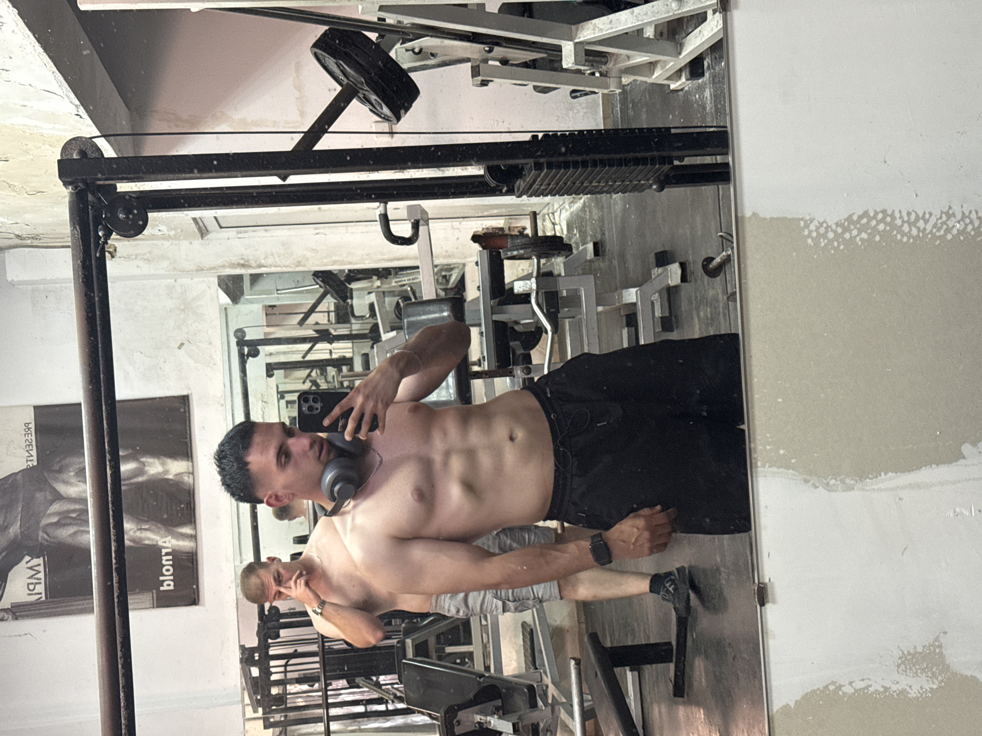 In a gym located in Shumen, Bulgaria, two shirtless men are reflected in a mirror. The man in the foreground is taking a selfie with his phone and wearing headphones around his neck. He is wearing black shorts. The man in the background is wearing grey cargo shorts and has his hand near his face. Various gym equipment, including weight machines and a pull-up bar, are visible in the background. The gym appears somewhat dated, with peeling paint on the walls.