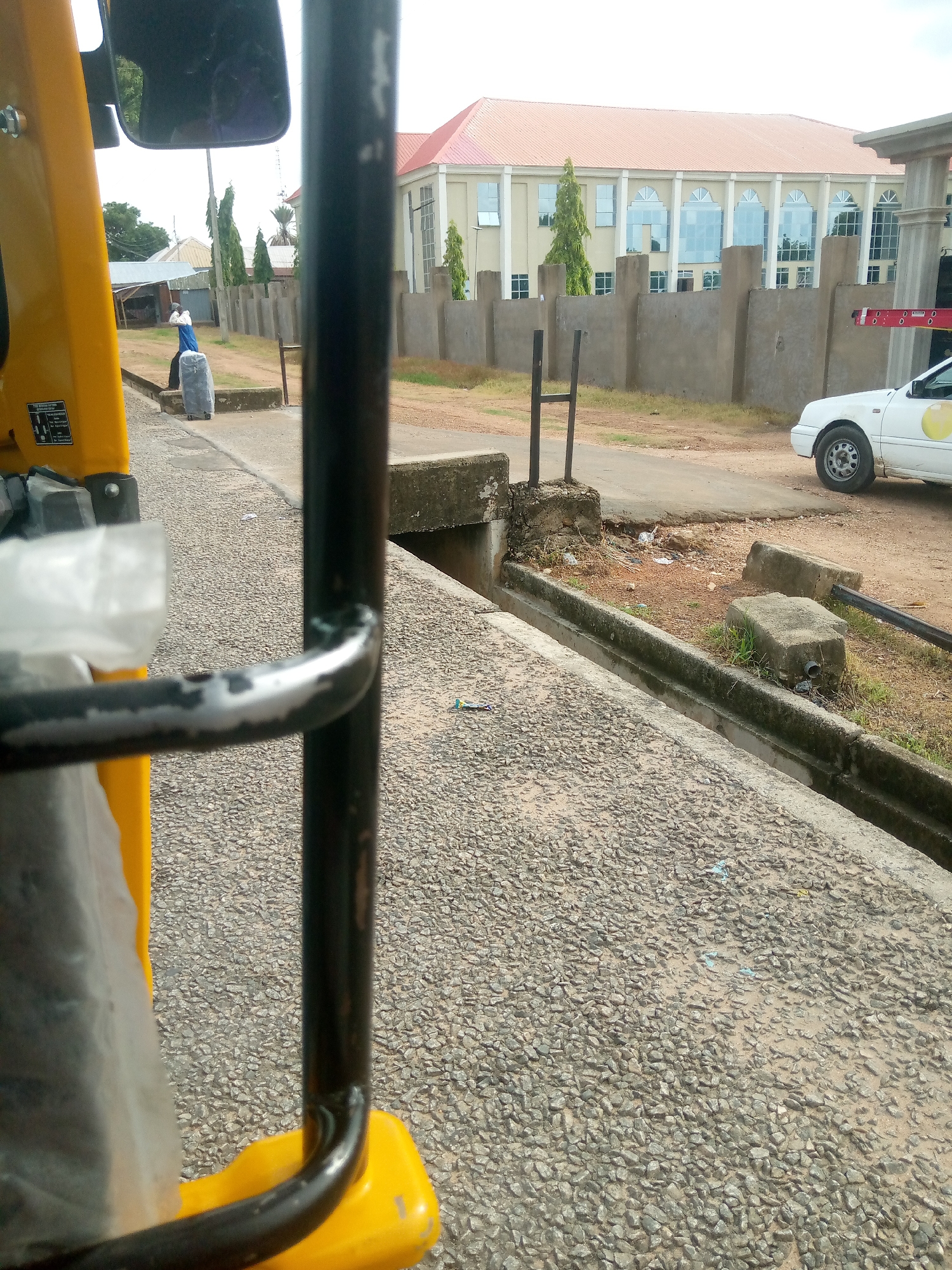 The image is taken from the perspective of someone inside a yellow, three-wheeled vehicle, looking outwards. In the foreground, a black metal bar, part of the vehicle's structure, is prominent. To the left, a large gray object wrapped in plastic is visible, also part of the vehicle's contents. The ground is covered in small, rough gravel.
In the middle ground, a person wearing a blue garment is seen near a concrete barrier, appearing to be moving a large gray suitcase. Beyond the person, a series of concrete walls line the area.
In the background, a large building with a reddish-orange roof and cream-colored walls is visible. The building has numerous windows, some of which appear to be reflective. Several tall, slender green trees stand in front of the building. A white car is parked to the right of the frame. The overall environment suggests an outdoor urban setting. The presence of the three-wheeled vehicle is indicative of its use as transport, and the building in the background resembles a public or commercial establishment. The scene is consistent with an area in Jalingo, Nigeria, where such vehicles are common for transportation and buildings of this architectural style are found.