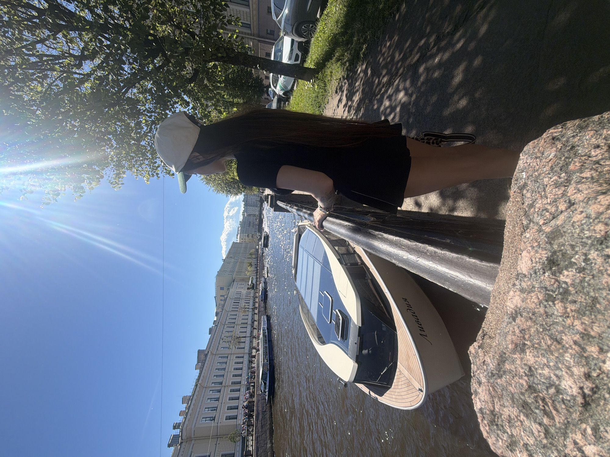 A young woman with long dark hair, wearing a black t-shirt dress and a light-colored baseball cap, stands on a sidewalk overlooking a canal in Saint Petersburg, Russia. She is facing away from the camera, looking towards the canal. In the canal, a modern-looking beige and black boat with a solar panel roof is docked. The boat has the name "Ливадия" inscribed on its side in Cyrillic script. Across the canal, historic cream-colored buildings with arched windows line the embankment. The sun is shining brightly, casting shadows from trees onto the sidewalk. Cars are parked on the street behind the woman. The canal water is murky brown. The overall scene depicts a sunny day in the city, with the woman enjoying the view of the waterway and the architecture.