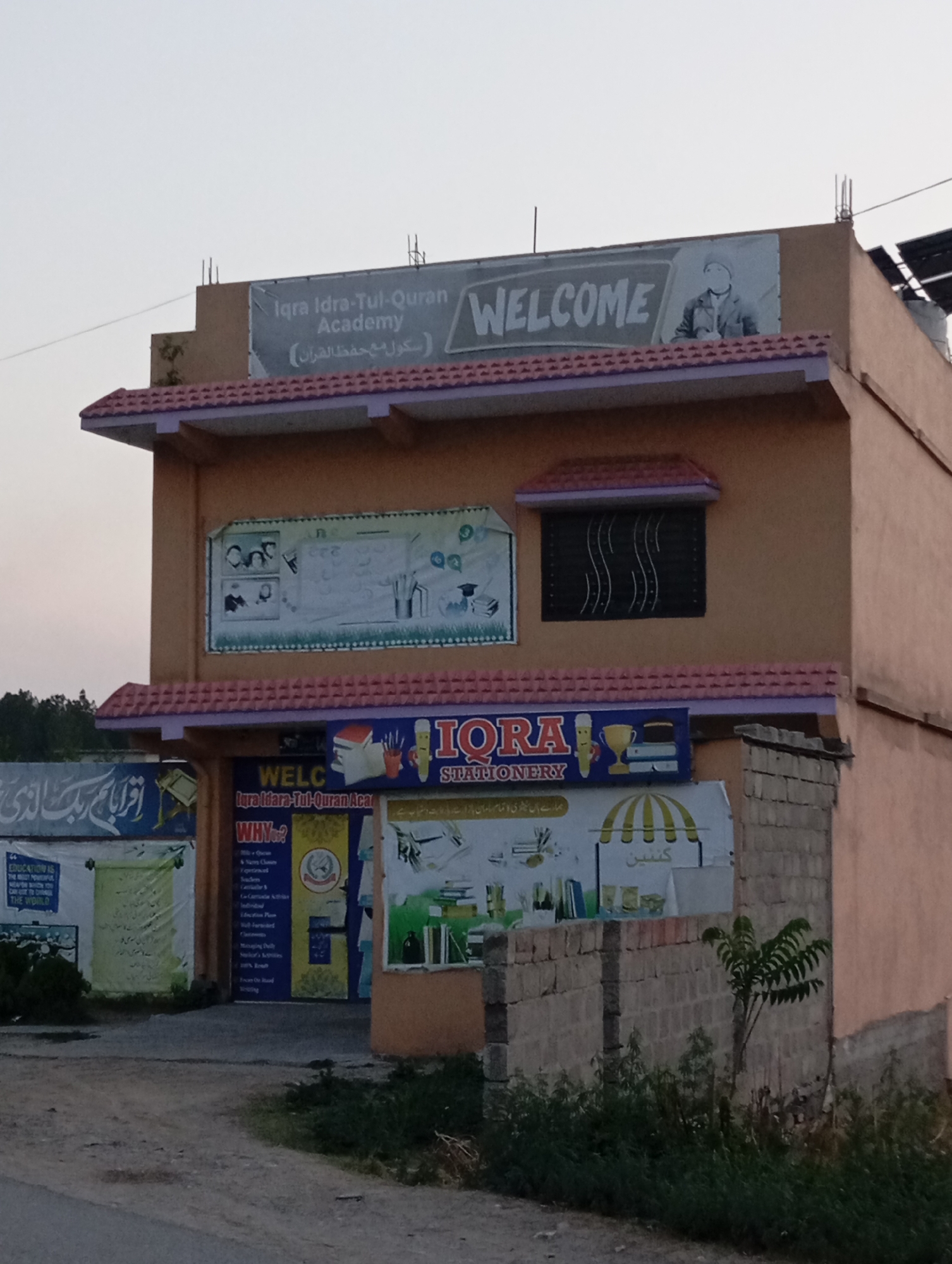 The image shows a two-story building with an orange facade and a red tiled roof, situated in Lassan, Pakistan. The building houses "Iqra Idra-Tul-Quran Academy" and "IQRA STATIONERY". A banner above the entrance reads "WELCOME". There are various signs on the building with text in English and possibly Urdu. The surrounding environment appears to be a street with some vegetation and a brick wall. The sky is overcast, suggesting it might be late afternoon or early evening. There are no people visible in the image.