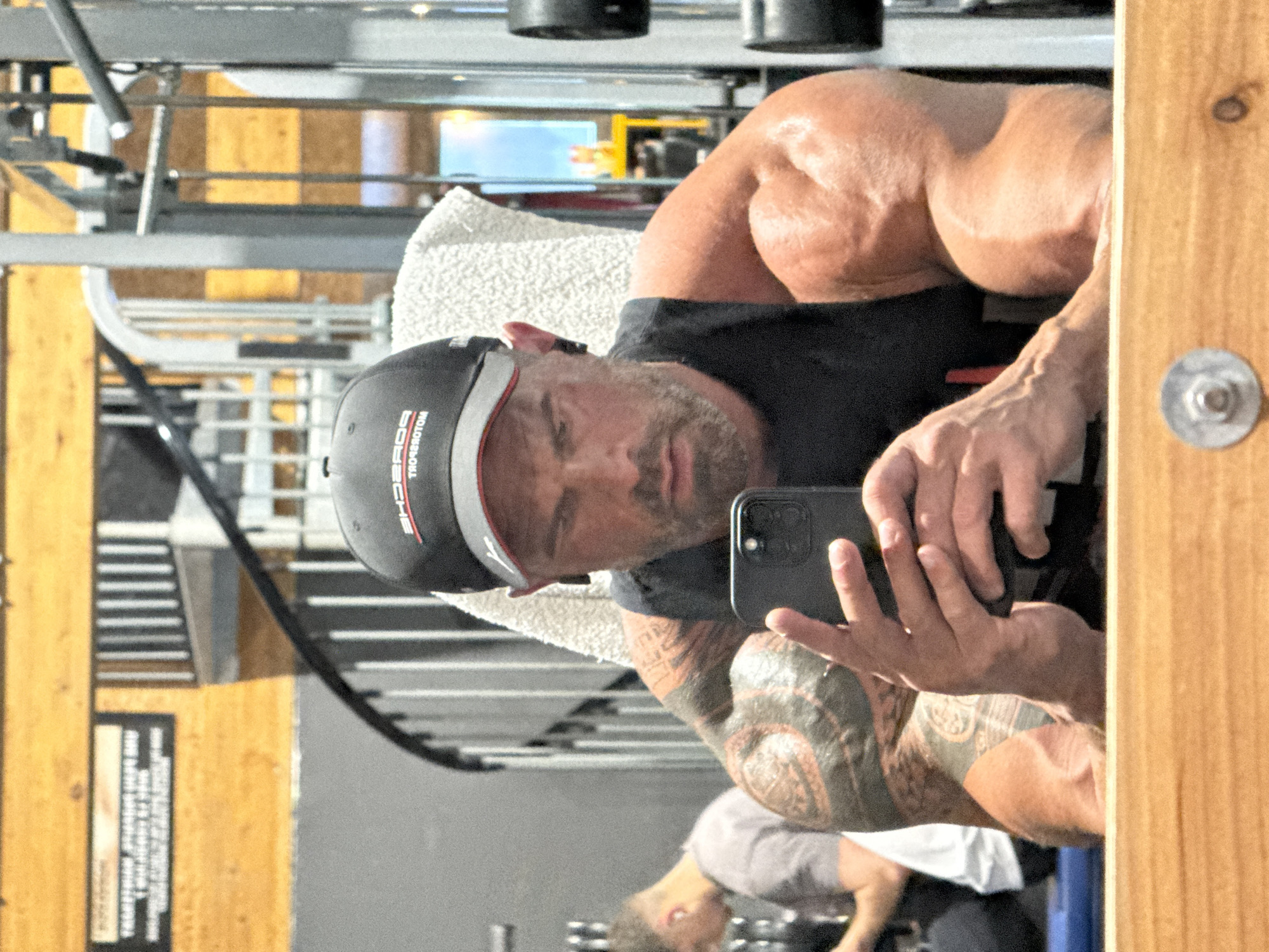 A man with a black and white cap is taking a selfie in a mirror inside what appears to be a gym. He has a gray beard and tattoos on his right arm. He is holding a black smartphone. The background shows gym equipment, a towel draped over a weight machine, and another person exercising in the background. Based on information included in the scene, the location is Jemelle, Belgium.