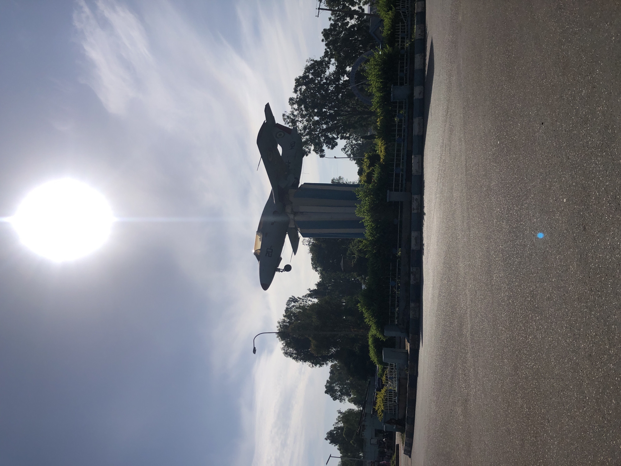 An outdoor, eye-level, wide shot captures a monument featuring a fighter jet in Jos, Nigeria, under a bright, sunny sky. The jet, angled slightly upward, is mounted on a tall, cylindrical pedestal striped with blue and white. The pedestal itself is surrounded by green bushes and a low, decorative fence. In the foreground, a paved road dominates the lower half of the frame. The background reveals trees and buildings, and a streetlamp is visible to the left of the monument. The sun is prominently positioned in the upper portion of the sky, causing lens flare. The overall impression is a clear day in an urban setting.