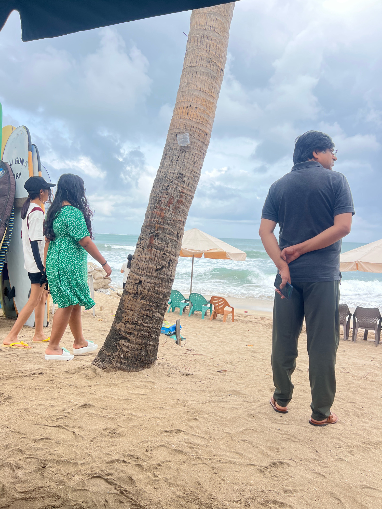 The image shows a beach scene in Kuta, Indonesia, on a cloudy day. In the foreground, a man in a dark grey polo shirt and dark green pants stands with his back to the camera, holding a dark-colored phone in his right hand behind his back. To his left, two young women are walking away from the camera. The woman on the left wears a white t-shirt with black sleeves and shorts, a black baseball cap, and yellow flip-flops. The woman on the right wears a bright green floral-patterned dress and white slip-on shoes. They are walking on a sandy beach littered with dried leaves and debris.
Behind the people, a large, rough-barked palm tree trunk dominates the center of the image, leaning slightly towards the left. In the background, the ocean is visible with moderate waves, and beach umbrellas and chairs are set up on the sand. Several surfboards are leaning against a structure on the far left side of the image. The sky is overcast with dark clouds, creating a subdued lighting condition.