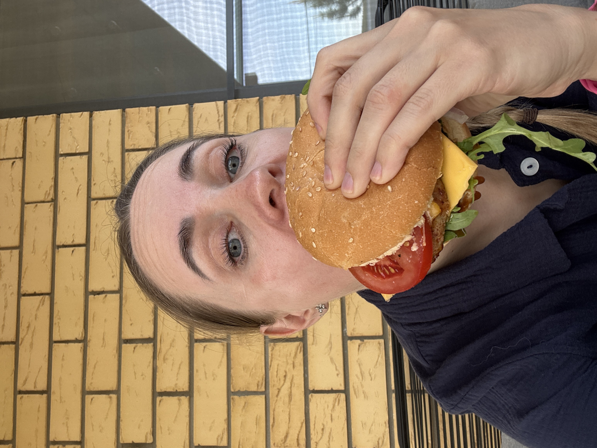 A woman with fair skin and blue eyes is holding a hamburger in front of her face. The hamburger appears to contain a slice of tomato, cheese, lettuce, and a meat patty. She is wearing a dark blue collared shirt with a visible button. The background shows a brick wall and a window. The location is suggested as Chaikovsky, Russia.